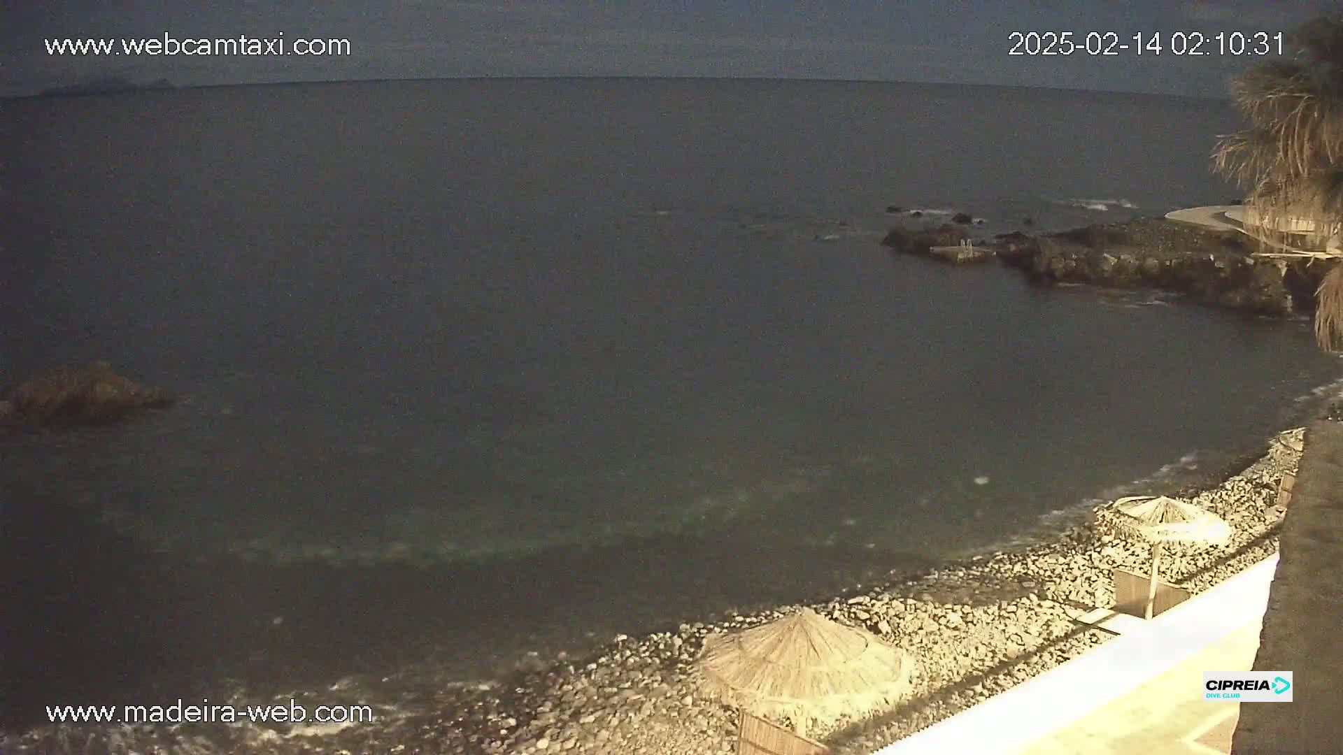 A dark, nighttime view of a rocky shoreline with calm water and a few beach umbrellas.