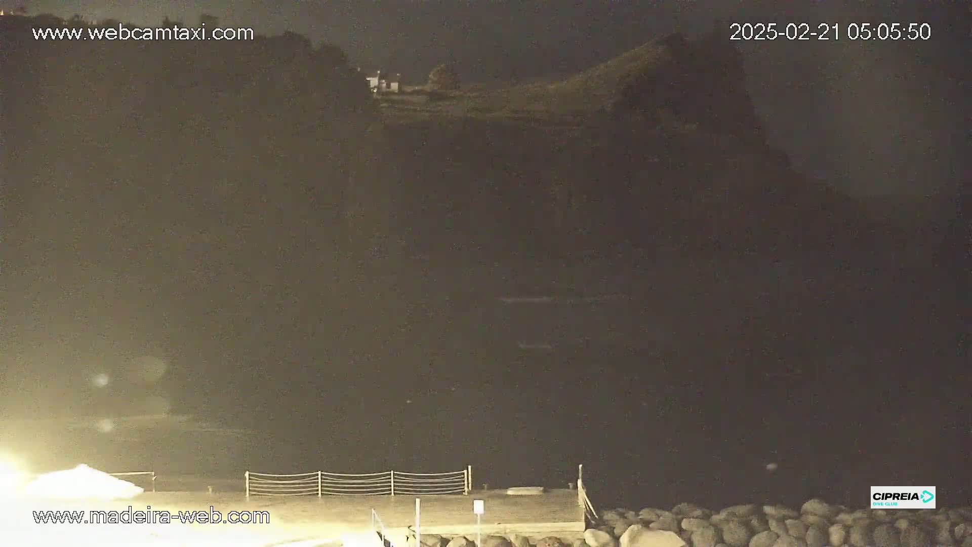 A dark, nighttime coastal scene shows a rocky cliff and calm water, with a small structure visible on the clifftop.