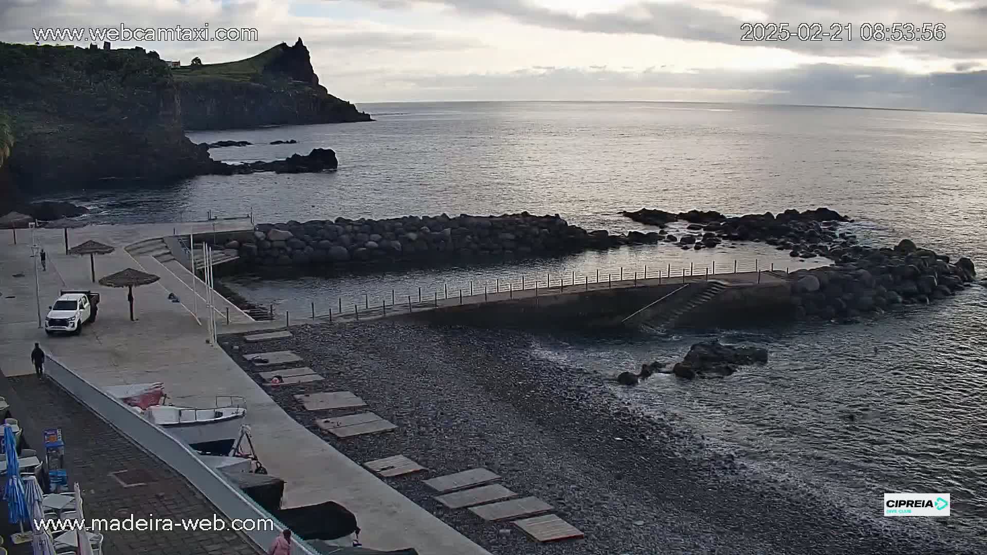 A rocky coastline with a small harbor, a pebble beach, and a calm ocean under a cloudy sky.