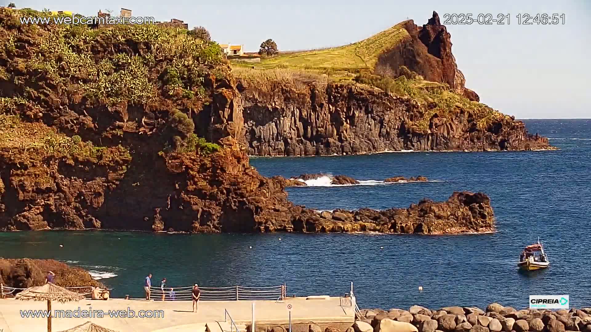 A rocky, dark-brown coastline with a small boat in the ocean on a sunny day.