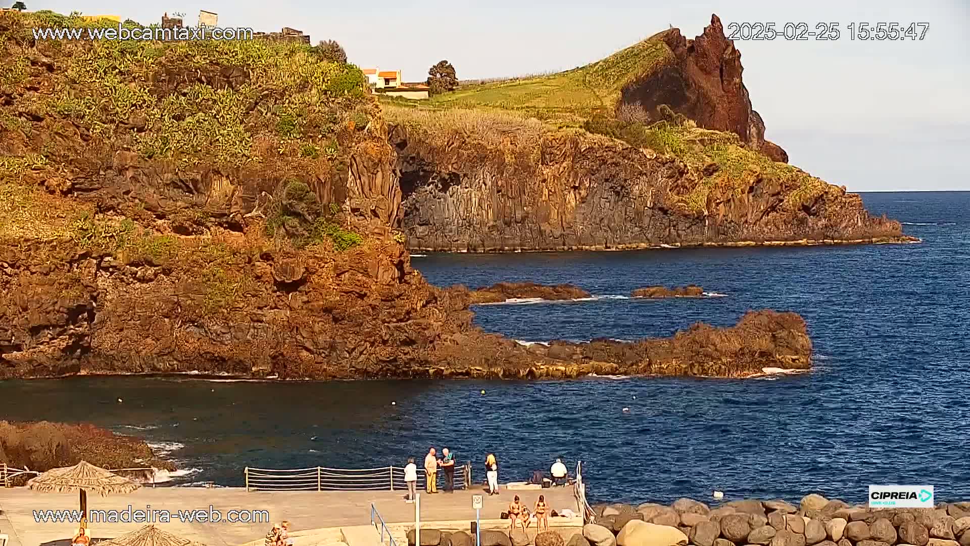 A rocky coastline with dark blue water under sunny skies, featuring several people standing on a small dock near a calm, sheltered cove.