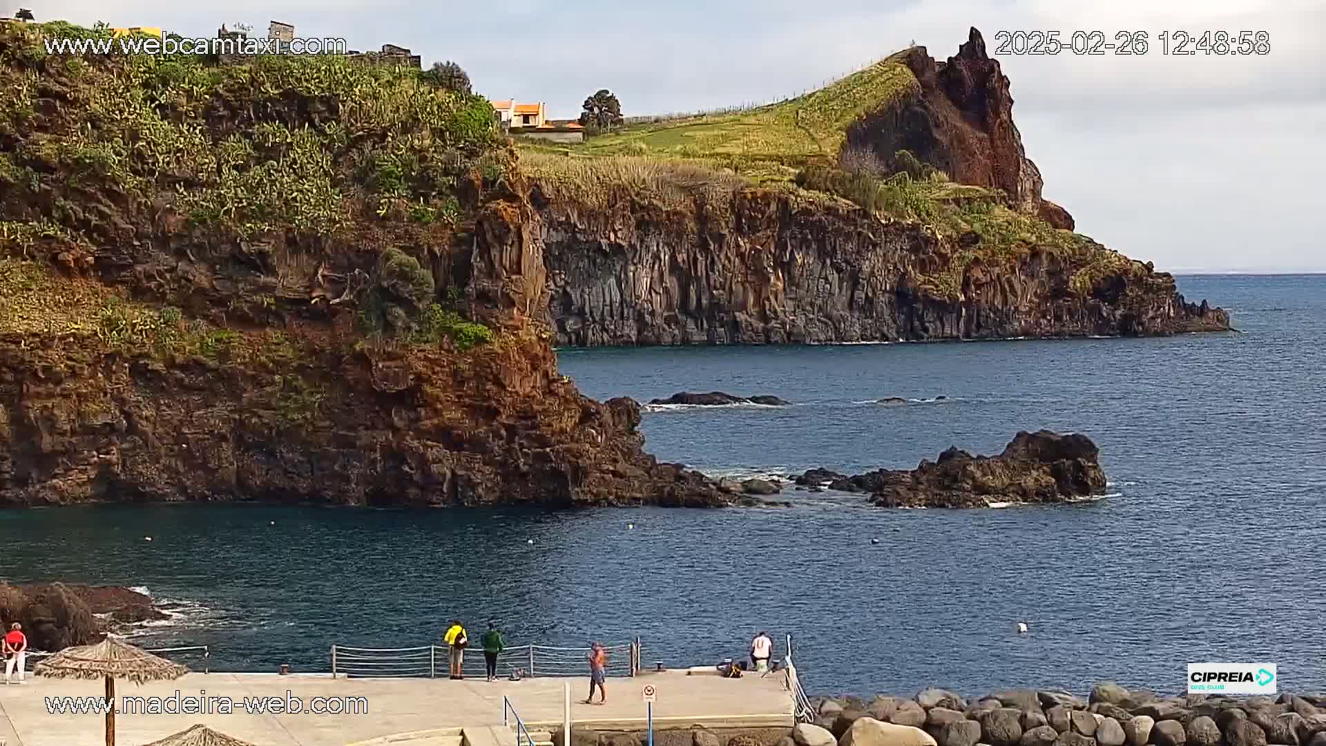 A rocky coast with a calm ocean, a small pier with several people, and a hillside with vegetation in the background under a partly cloudy sky.