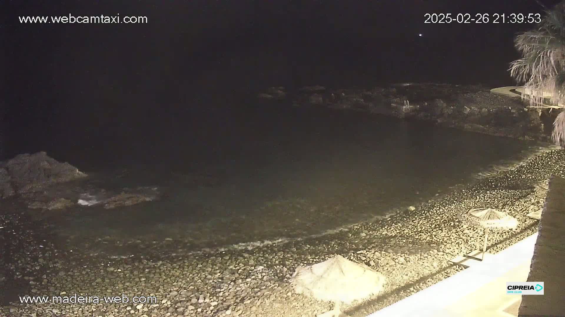 A dark, rocky beach at night with calm water lapping the shore, and a few closed umbrellas on the sand.