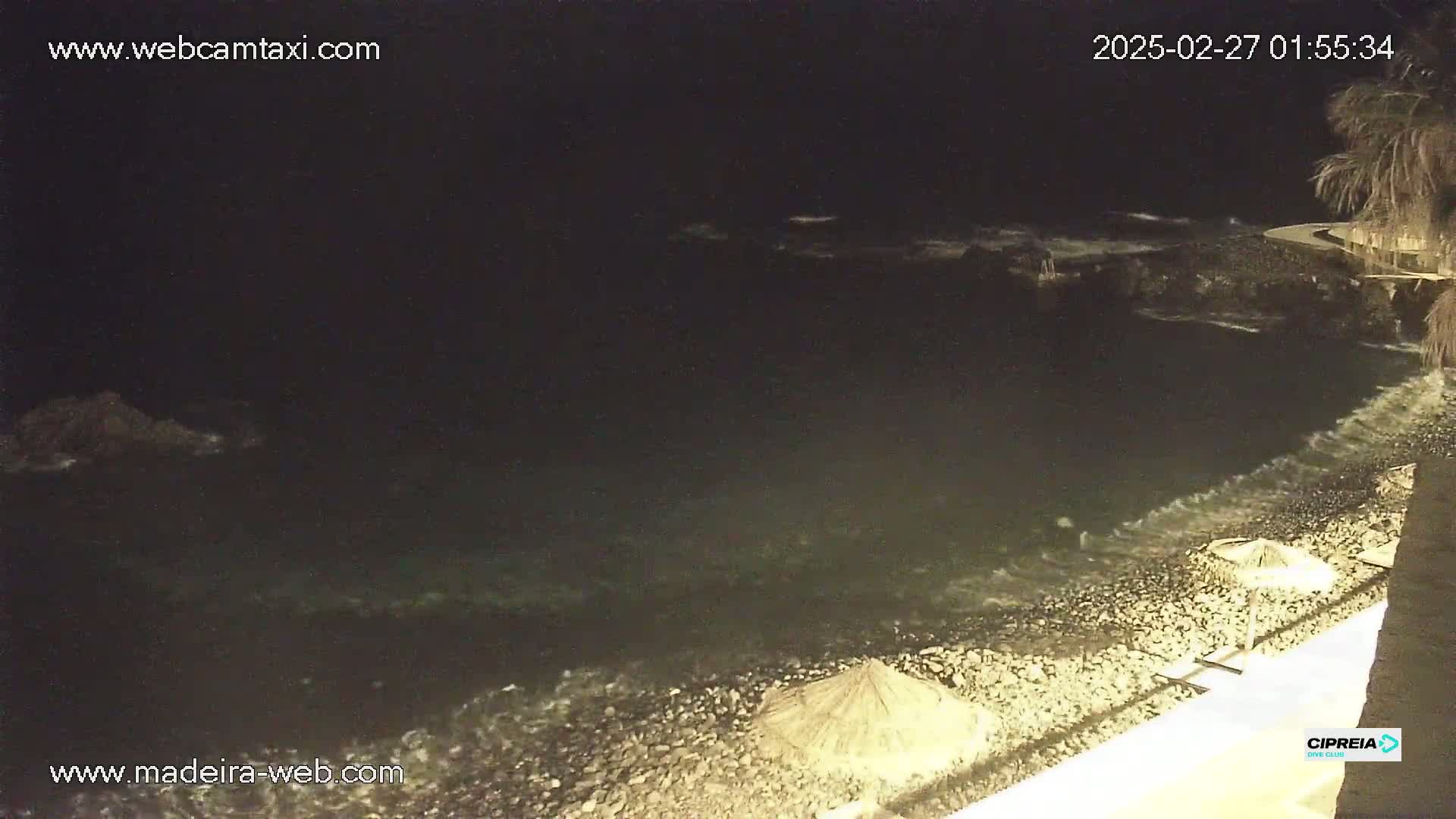 A dark, nighttime beach scene shows a rocky shore with gentle waves lapping at the sand, and a few closed beach umbrellas are visible in the foreground.