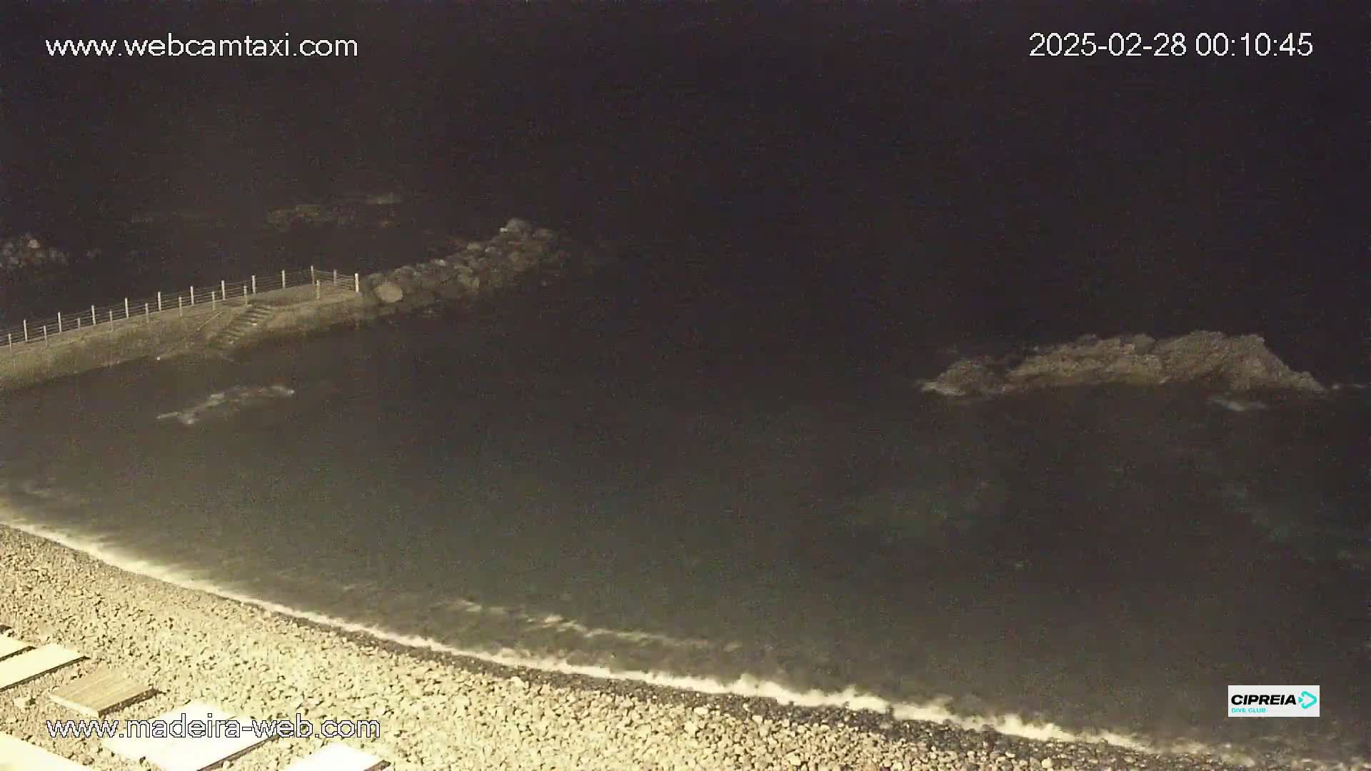 A dark, nighttime beach scene shows a rocky shoreline with gentle waves lapping at the shore.