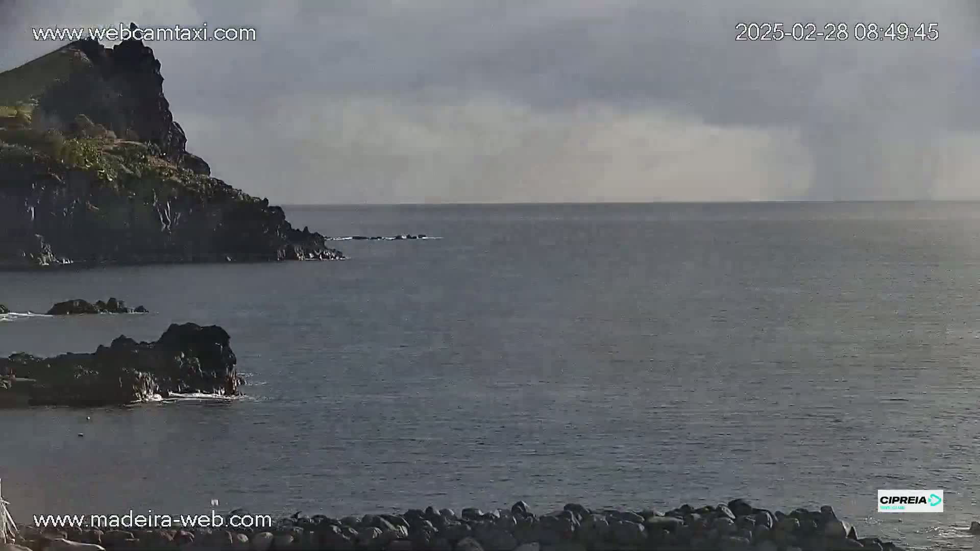 A dark, rocky coastline meets a calm, grey ocean under an overcast sky.