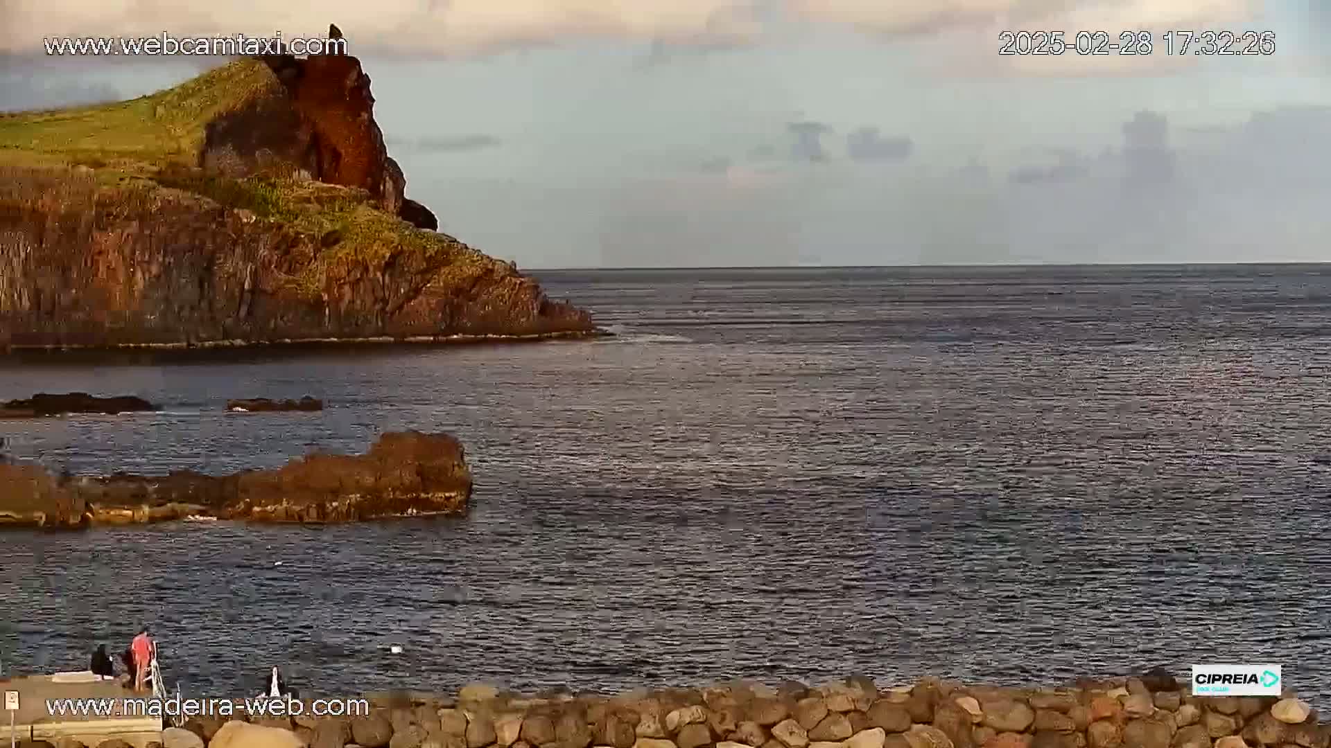 A rocky, dark-colored cliff overlooks a calm, dark-blue ocean under a mostly cloudy sky.