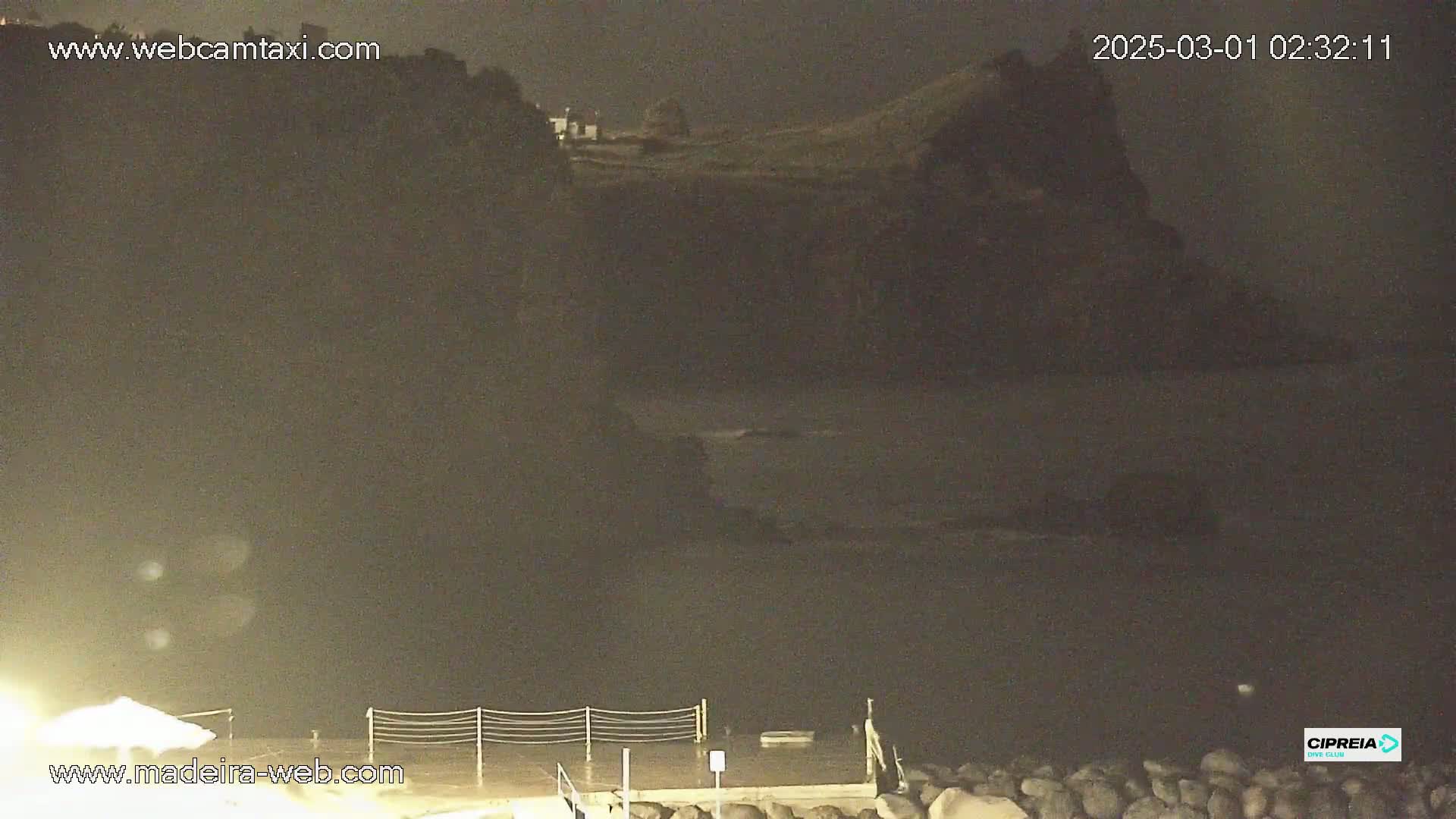 A nighttime view of a dark, rocky coastline and calm water under a dark sky.