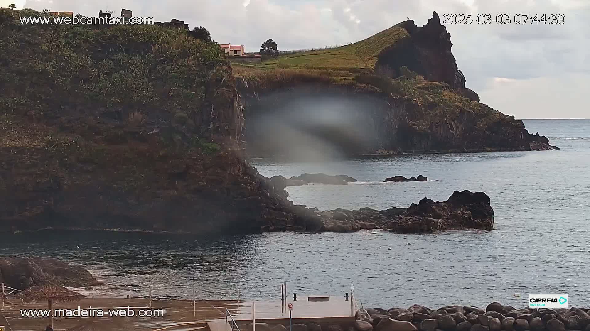 A rocky coastline with a small building on a cliff overlooking calm ocean water under a cloudy sky.