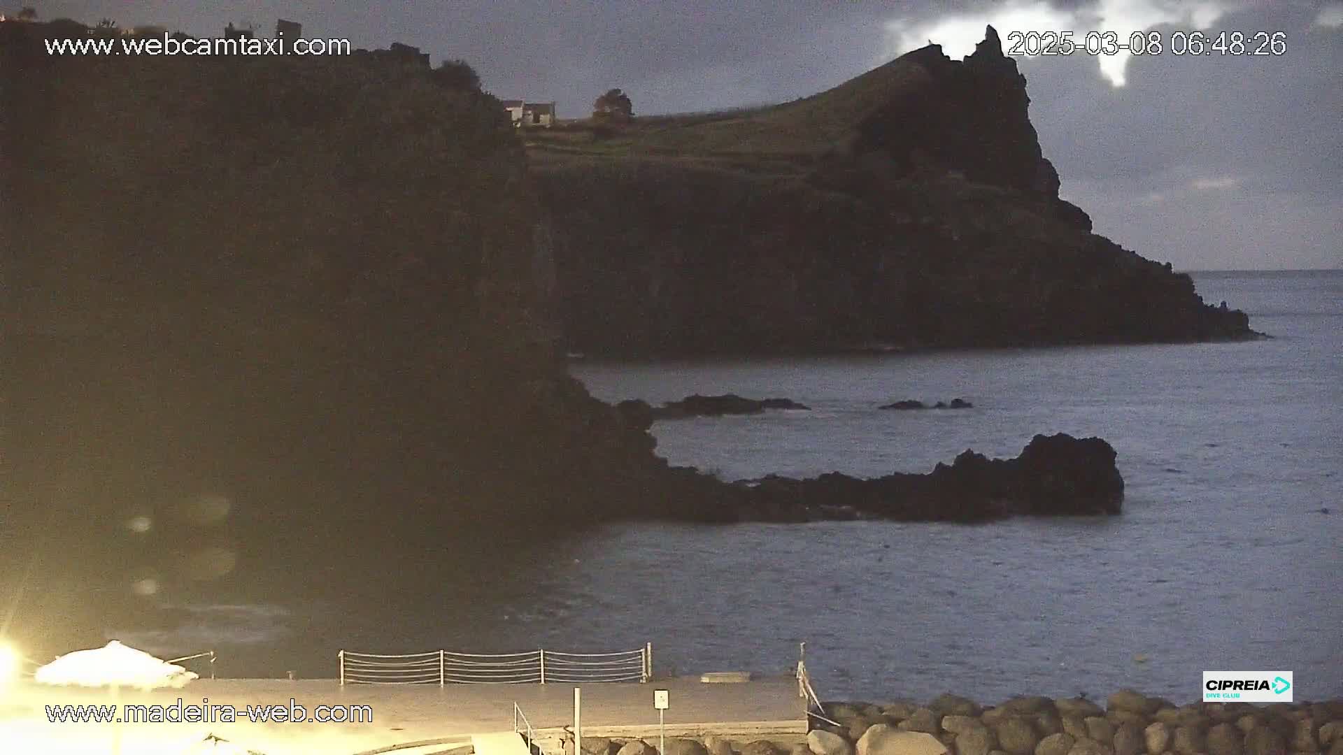 A dark, rocky coastline meets a calm, dark-blue ocean under an overcast sky at twilight.