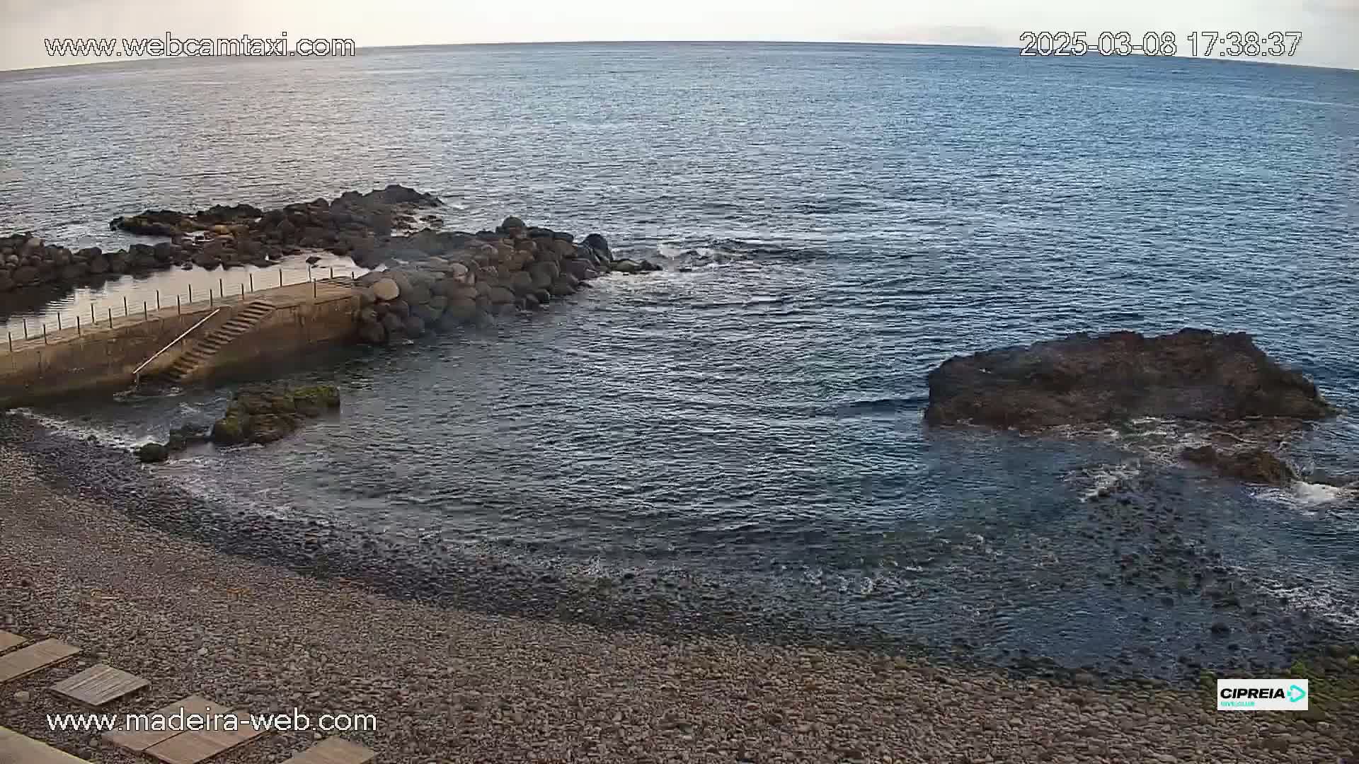 A rocky beach with a small concrete structure and steps leading into calm, dark-blue ocean water under a mostly clear sky.