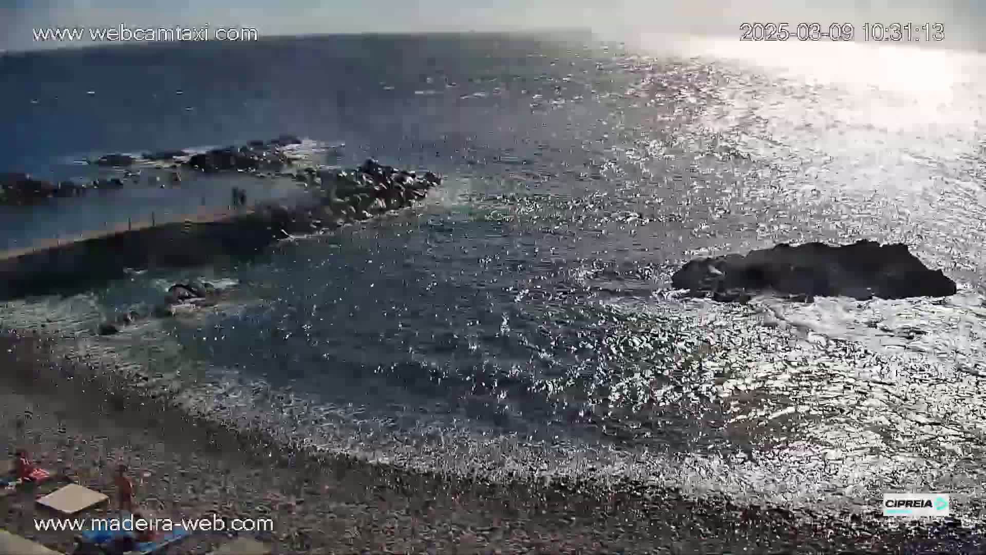 A rocky shoreline with a small man-made pool is partially visible, with a dark, wavy ocean reflecting sunlight under a mostly clear sky.