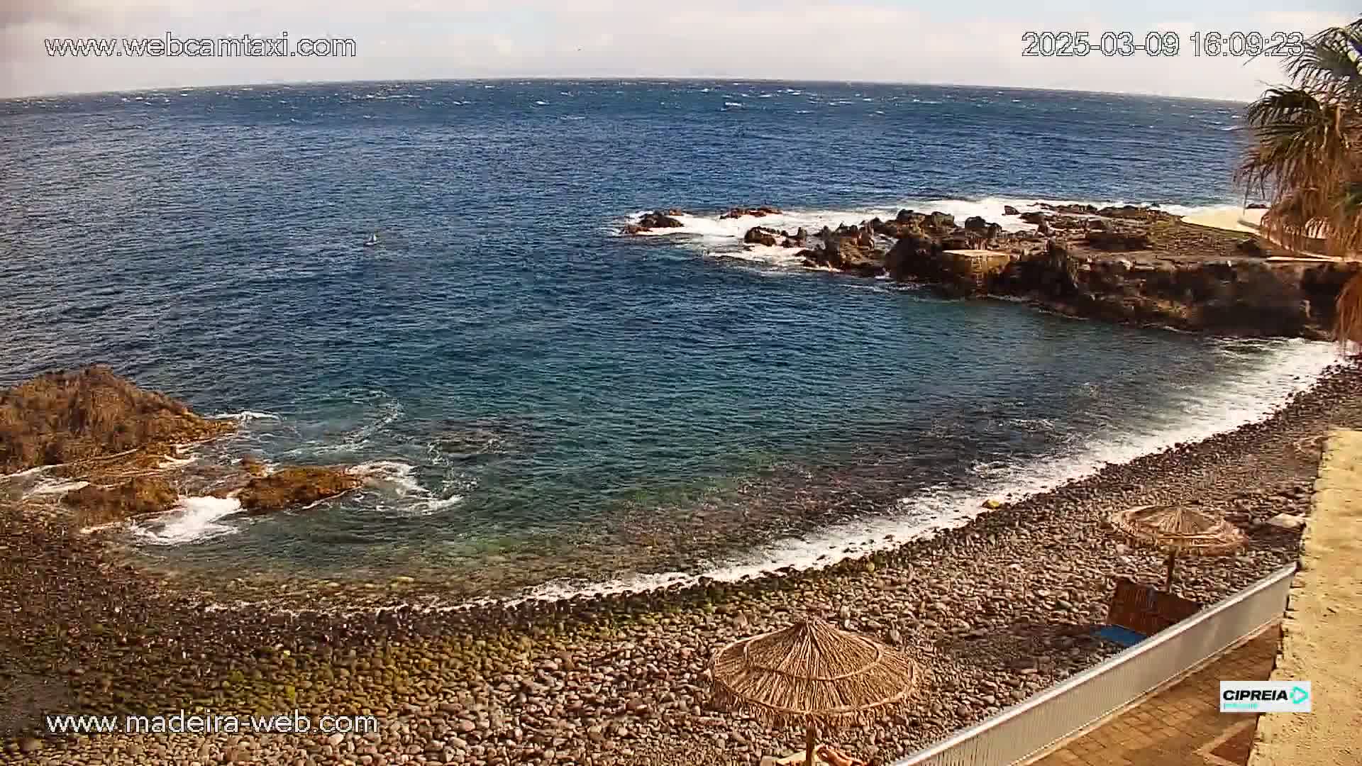 A rocky, pebble beach with calm, clear water laps against the shore under a partly cloudy sky.