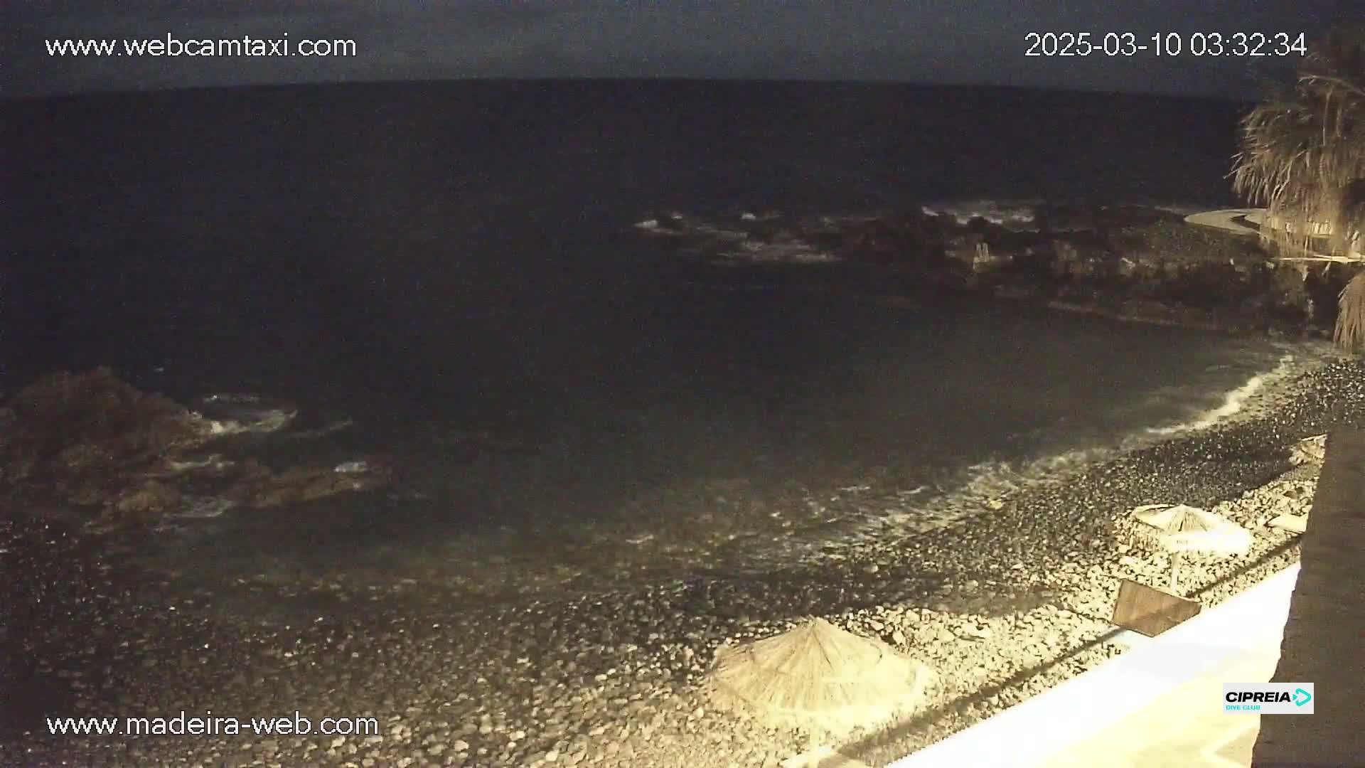 A dark, rocky beach at night with gentle waves lapping the shore, under a mostly cloudy sky.
