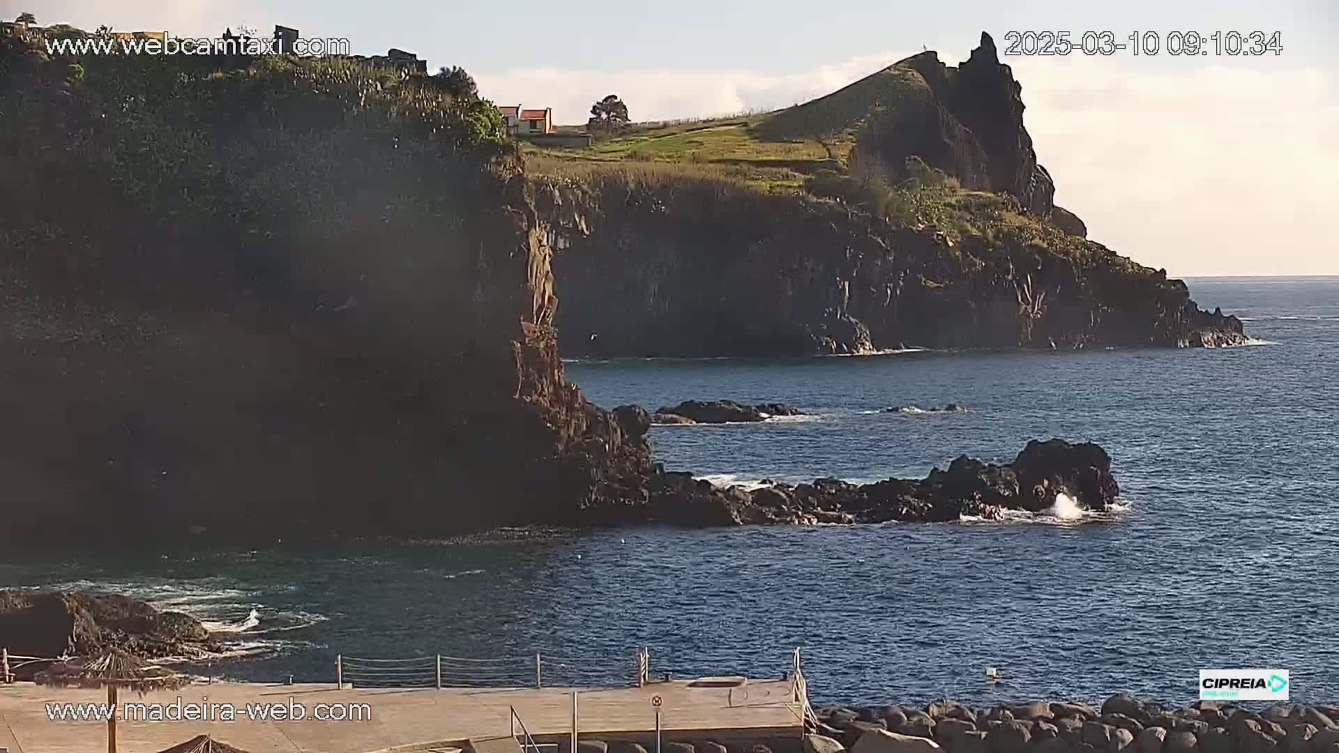 A rocky, dark-colored cliff overlooks a calm, blue ocean under a mostly clear sky.