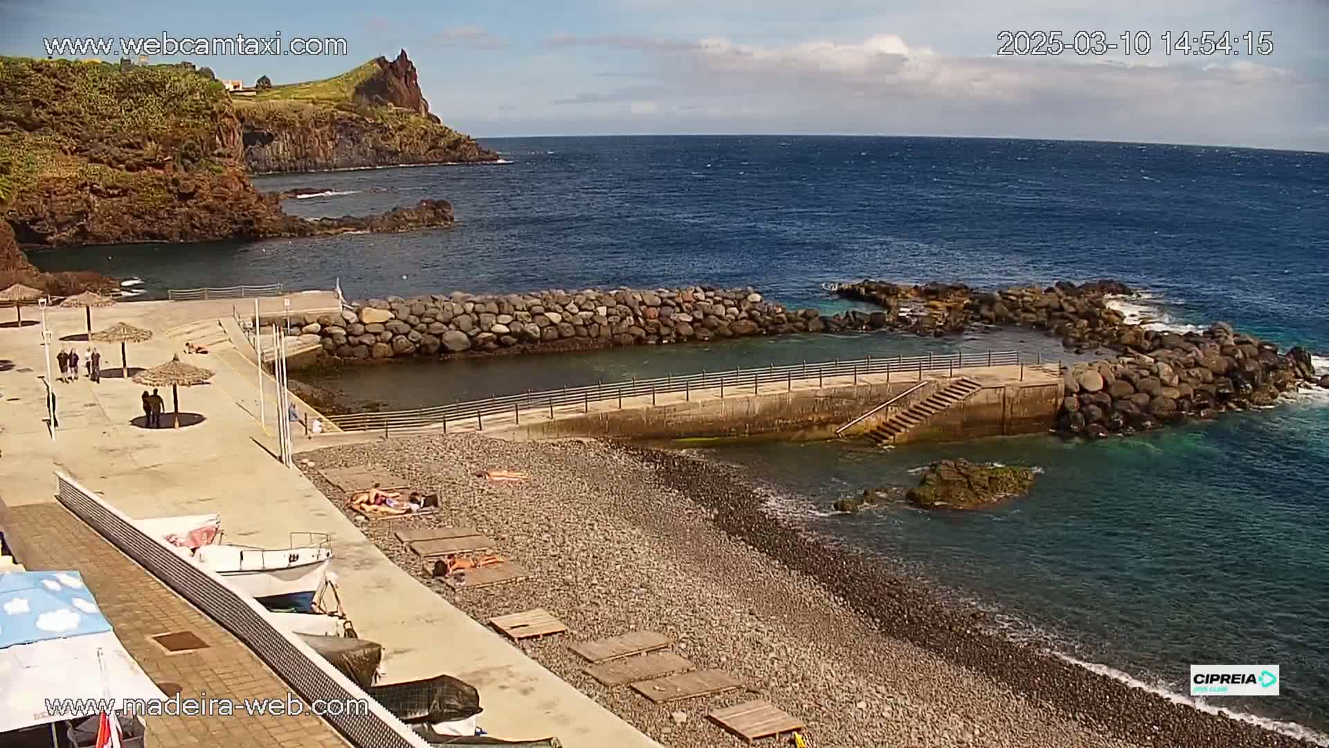 A rocky coast with a small pebble beach, a man-made harbor, and several people are relaxing under umbrellas on a sunny day.