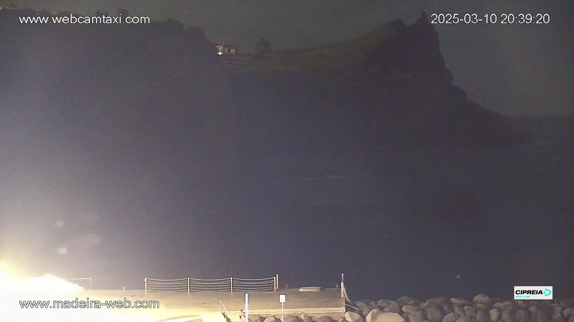 A dark, nighttime coastal scene shows a rocky cliff silhouetted against a dark sky, with a small structure visible near its top, and a low-lit dock in the foreground.