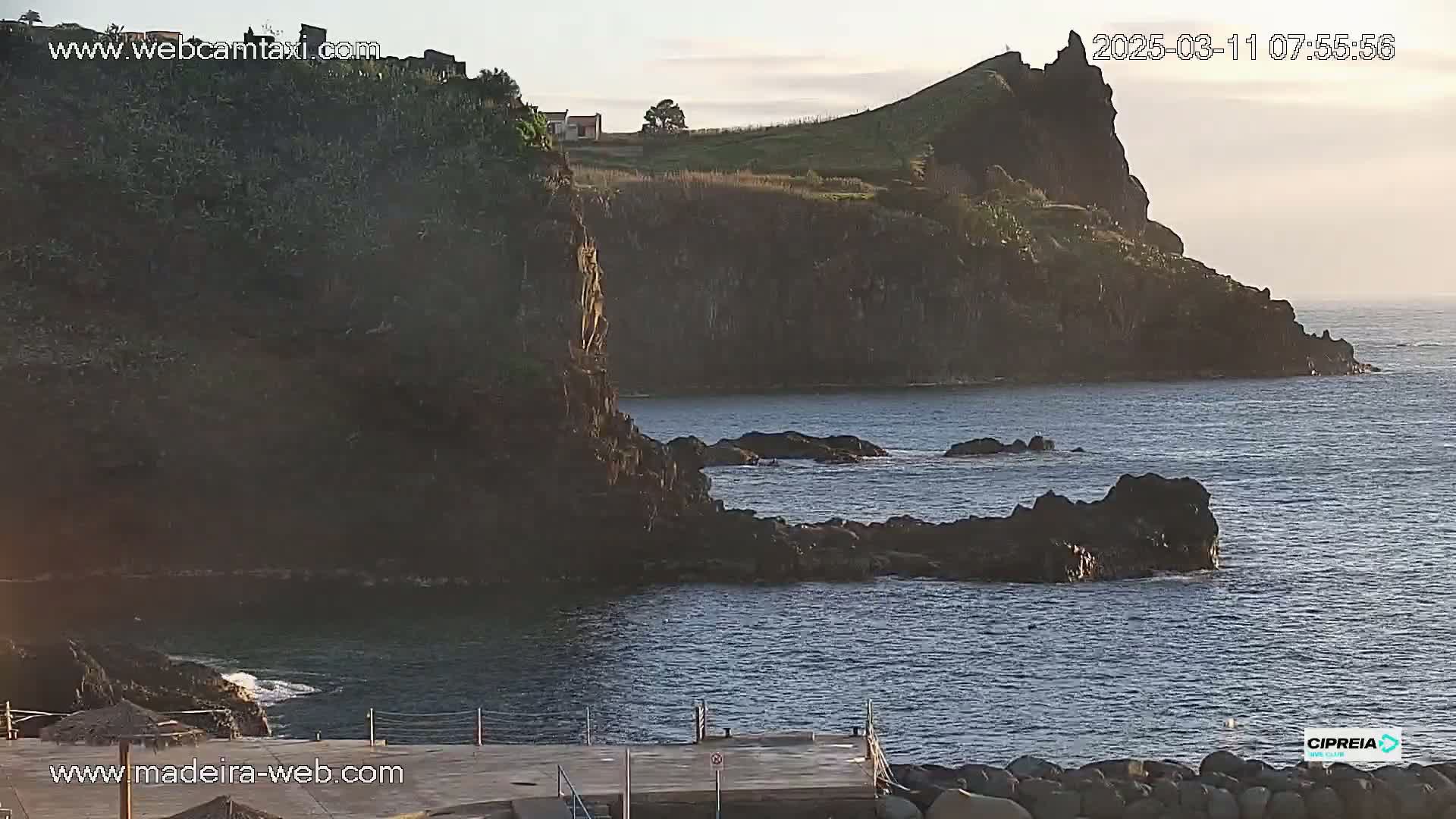 A rocky coastline with a small dock is visible at the bottom of a calm body of water under a mostly clear sky.