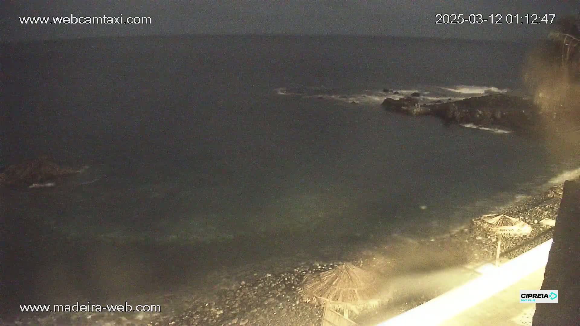A dark, nighttime coastal scene shows a rocky shoreline with small waves lapping against it, a few beach umbrellas, and a dark, somewhat stormy ocean beyond.