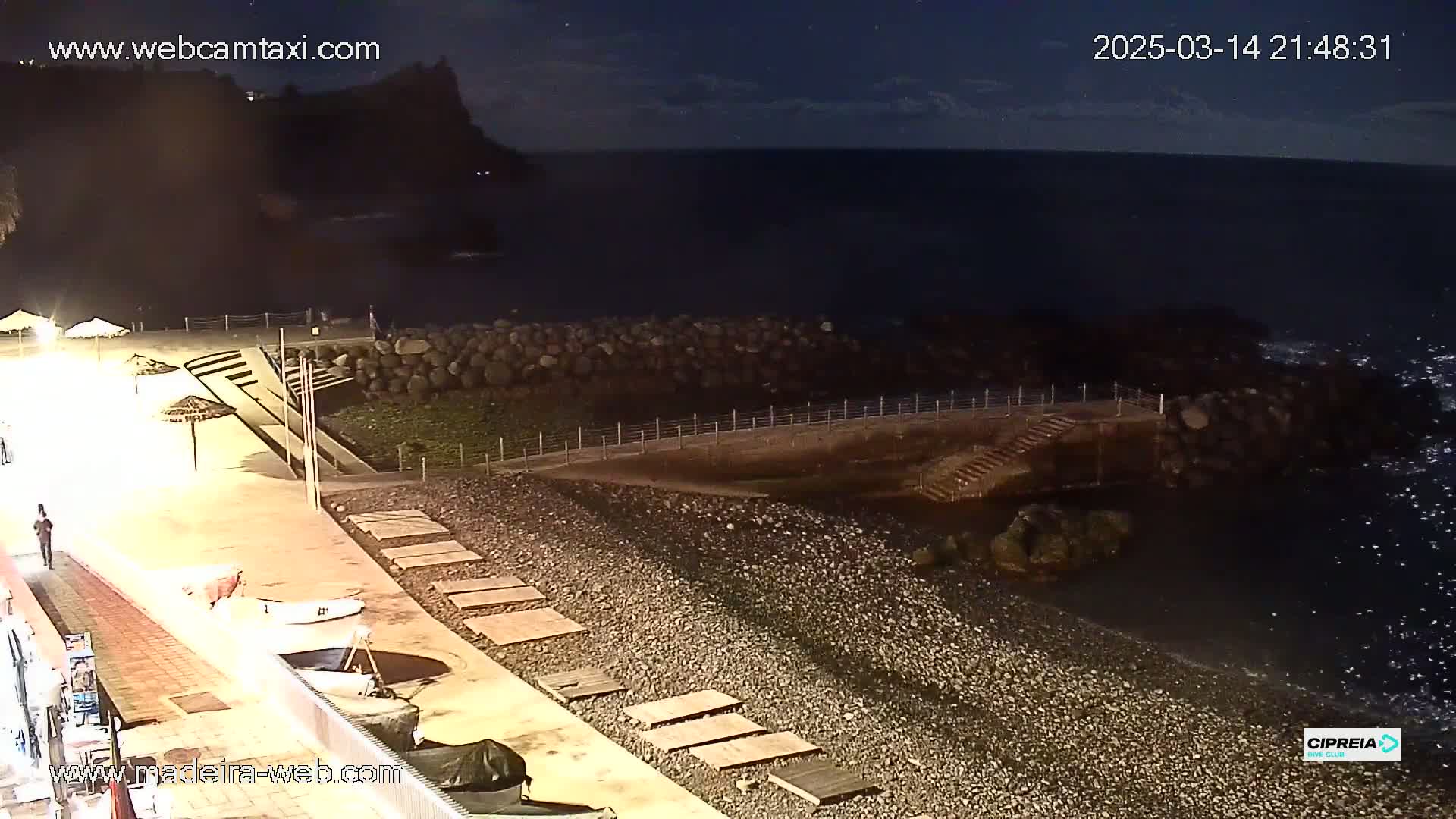 A nighttime view of a rocky beach with a calm dark ocean, a paved walkway, and several small boats pulled ashore under mostly clear skies.