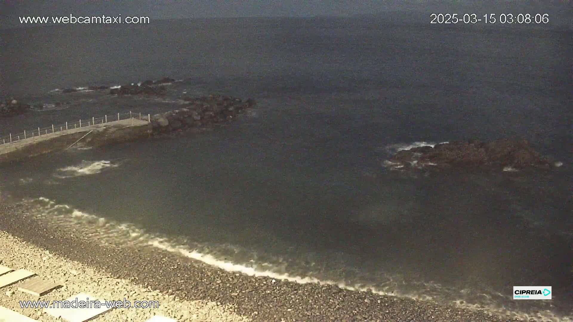 A rocky shoreline with a small jetty meets dark, somewhat choppy water under an overcast sky.