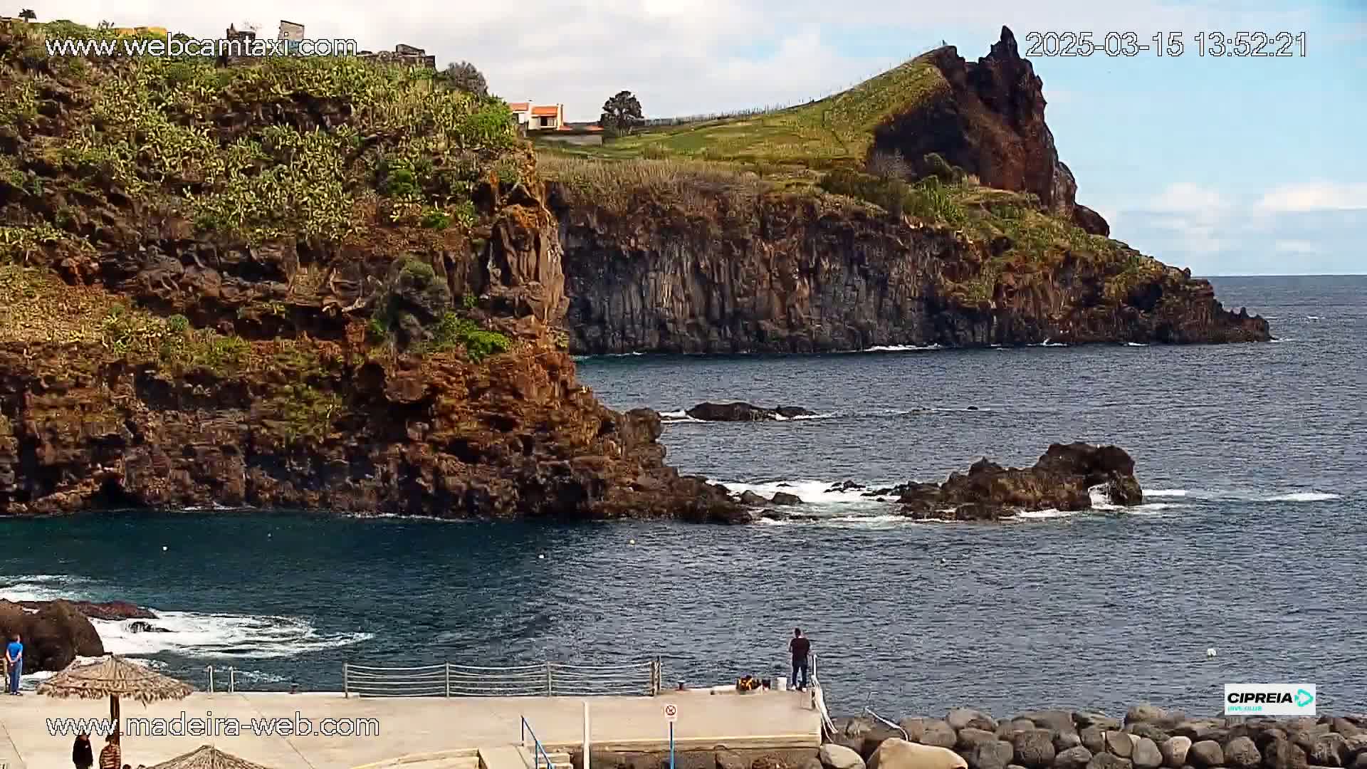 Dark, rocky cliffs meet a partially calm ocean, with a few people standing on a small jetty under partly cloudy skies.