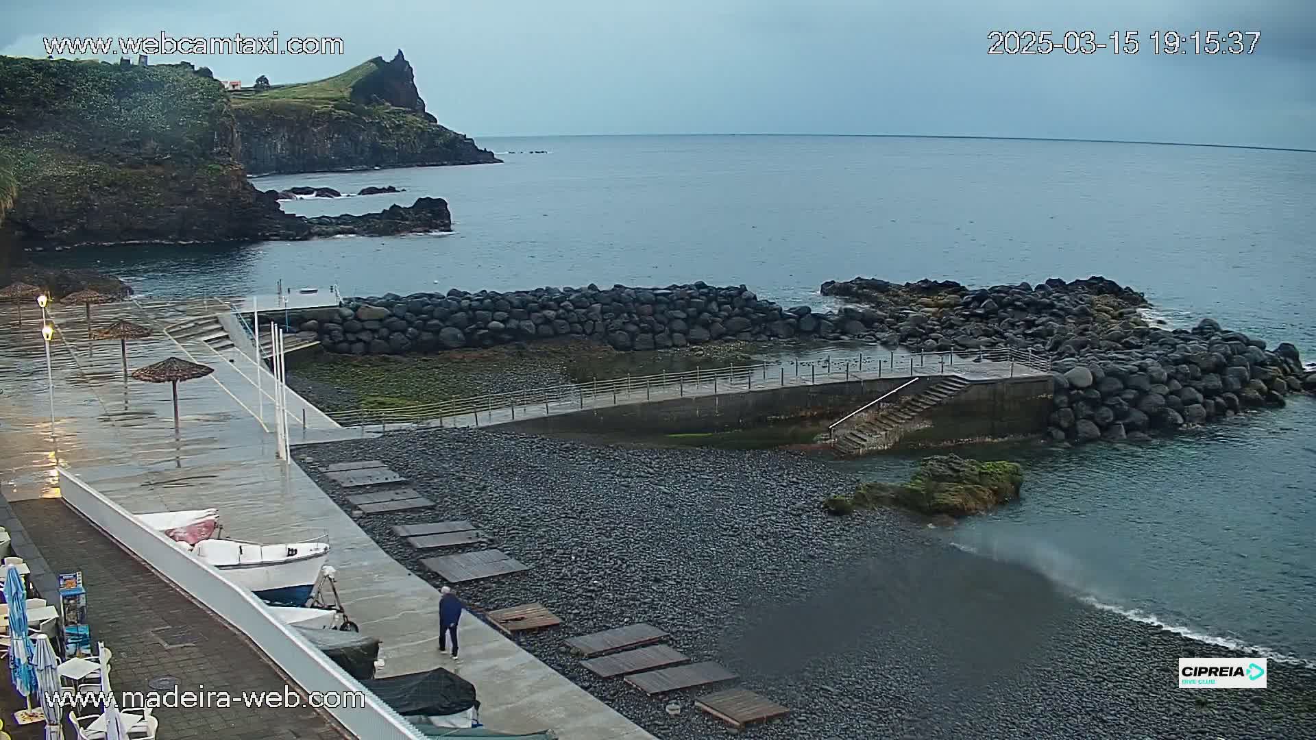 A wet, dark-colored pebble beach with a small harbor, a walkway, and a few boats is situated next to a rocky coastline and calm ocean under an overcast sky.