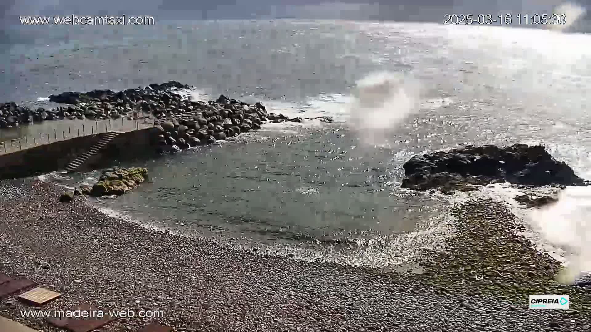 Waves crash against a rocky shoreline near a pebble beach with a stone staircase leading to the water.