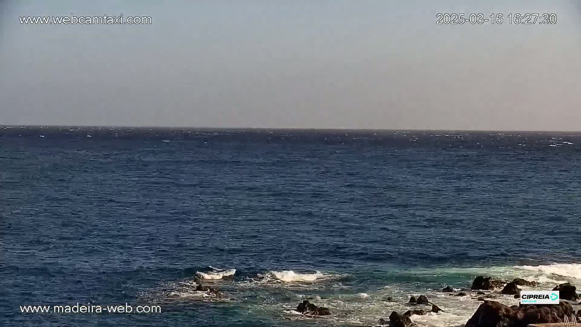 A mostly clear sky overlooks a dark blue ocean with small waves breaking against dark rocks in the foreground.