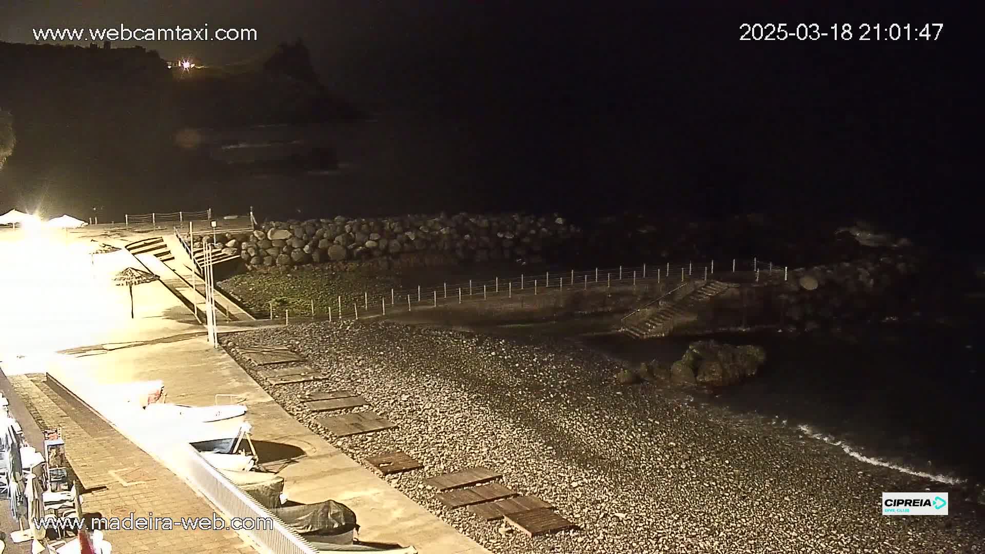 A dark, pebble beach at night is illuminated by artificial lights, with a low stone wall and some small boats visible near a paved walkway.