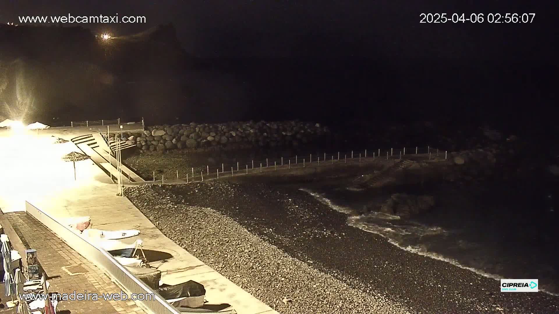 A nighttime view of a rocky shoreline with a paved walkway, small boats, and closed umbrellas, under dark, clear skies with gentle waves lapping the shore.