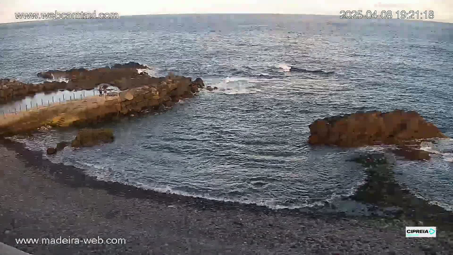 A rocky coastline with a small, dark-colored beach and calm ocean water under a mostly clear sky.