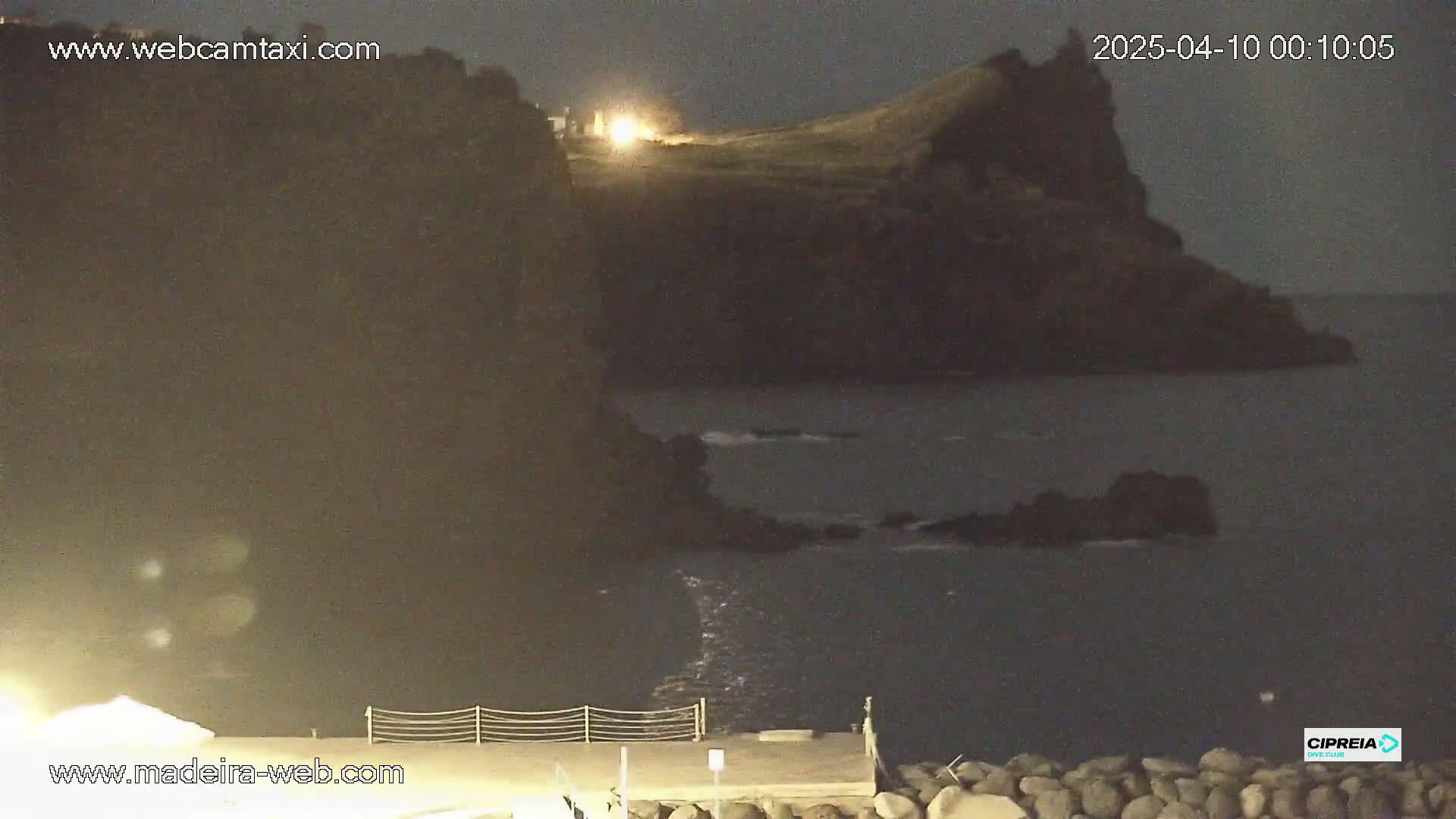 A nighttime coastal scene shows dark, rocky cliffs overlooking a calm, dark ocean under a clear, night sky; a light is visible atop one of the cliffs.