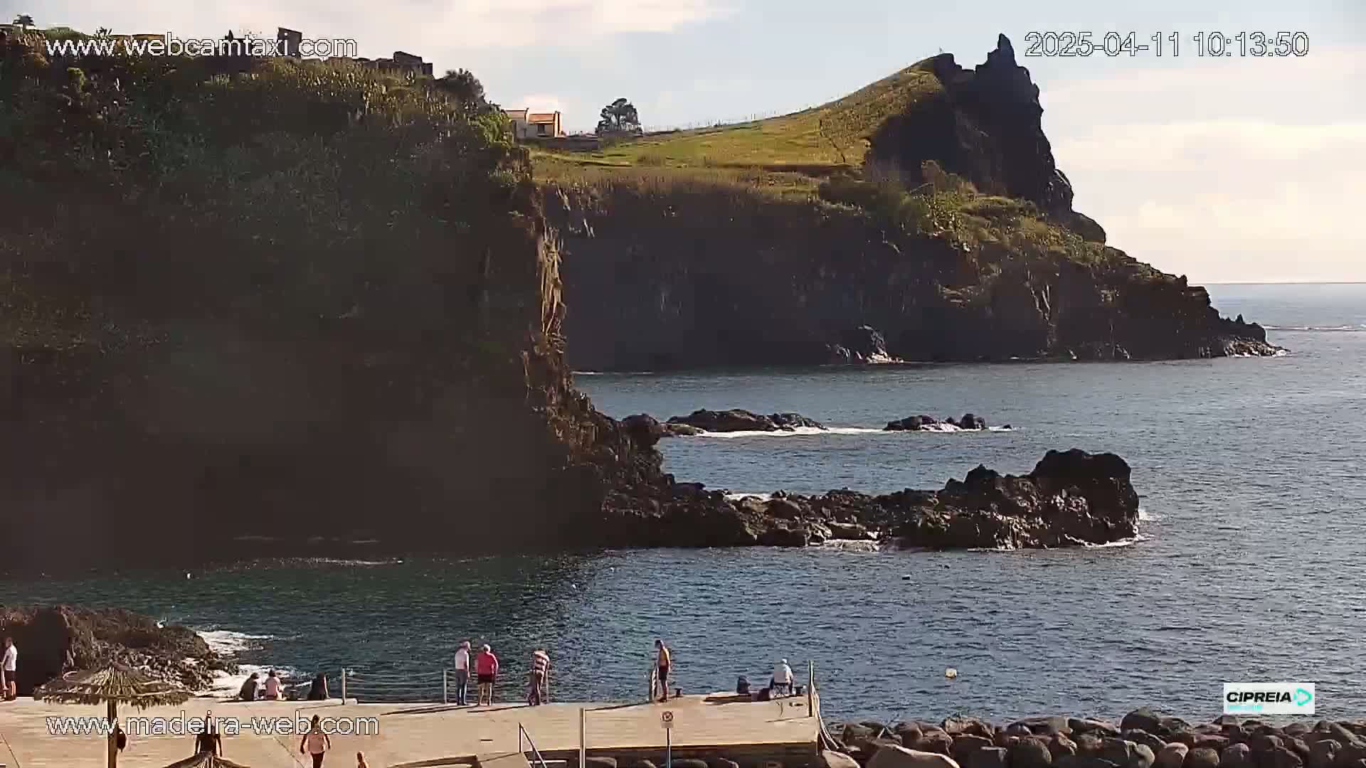 A rocky coastline with a few people standing on a dock overlooks calm ocean water under a partly cloudy sky.