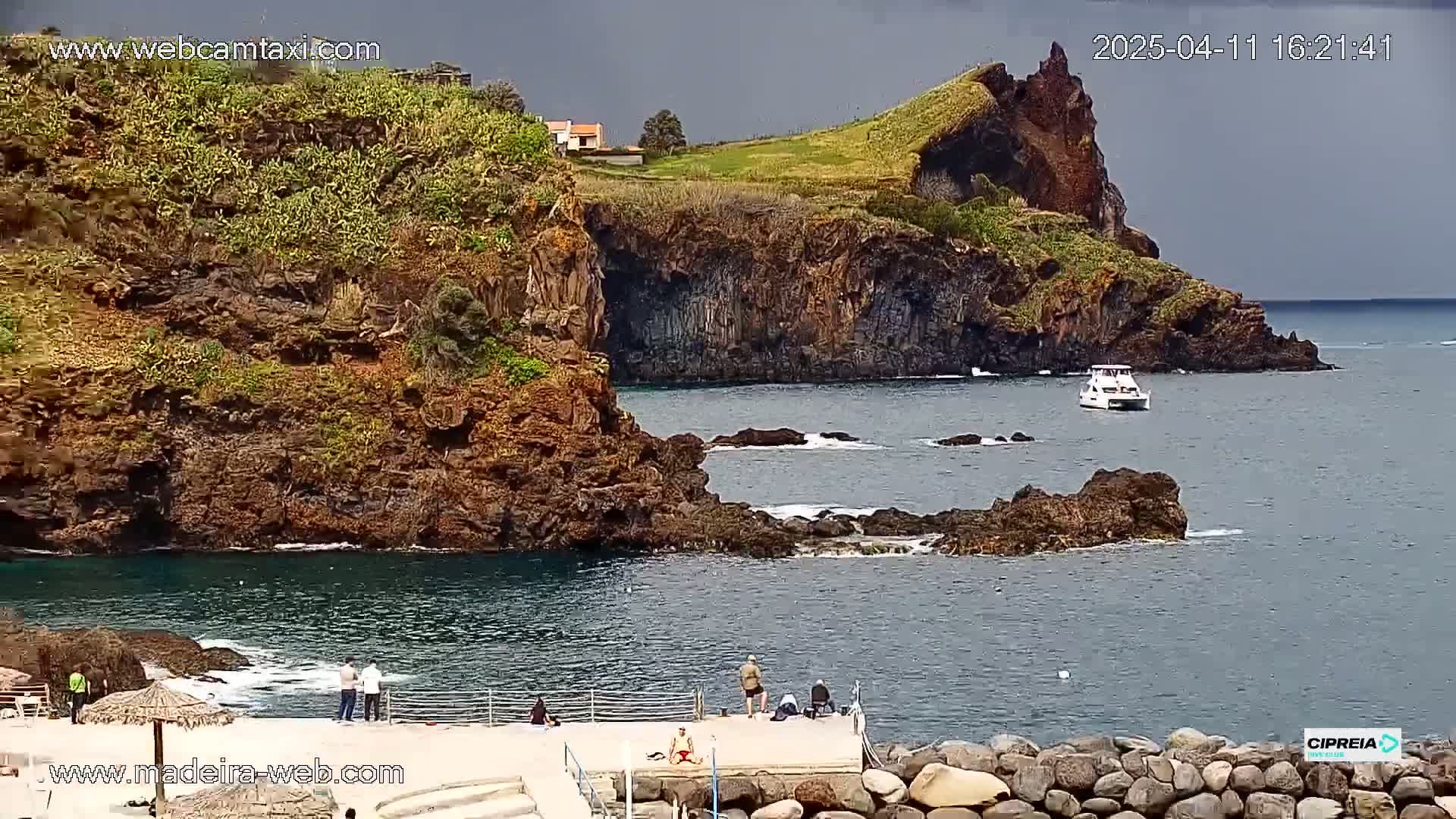 A rocky coastline with dark, volcanic-looking cliffs, a small boat in the water, and a few people standing on a concrete platform under an overcast sky.