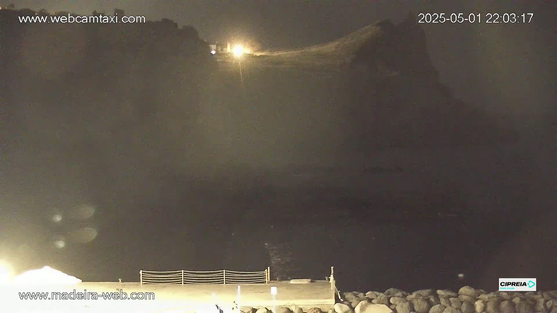 A nighttime coastal scene shows a dark, hazy body of water with a rocky shoreline and a distant, dimly lit cliff in the background.