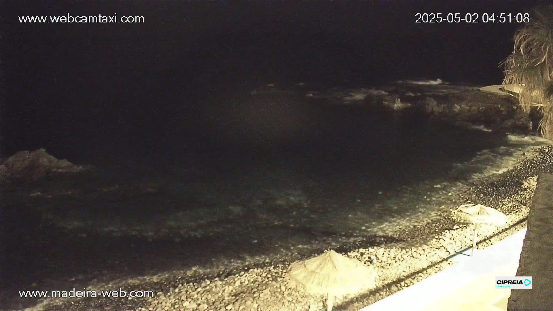 A dark, nighttime beach scene shows gently lapping waves on a pebble shore, with a partially visible thatched umbrella.