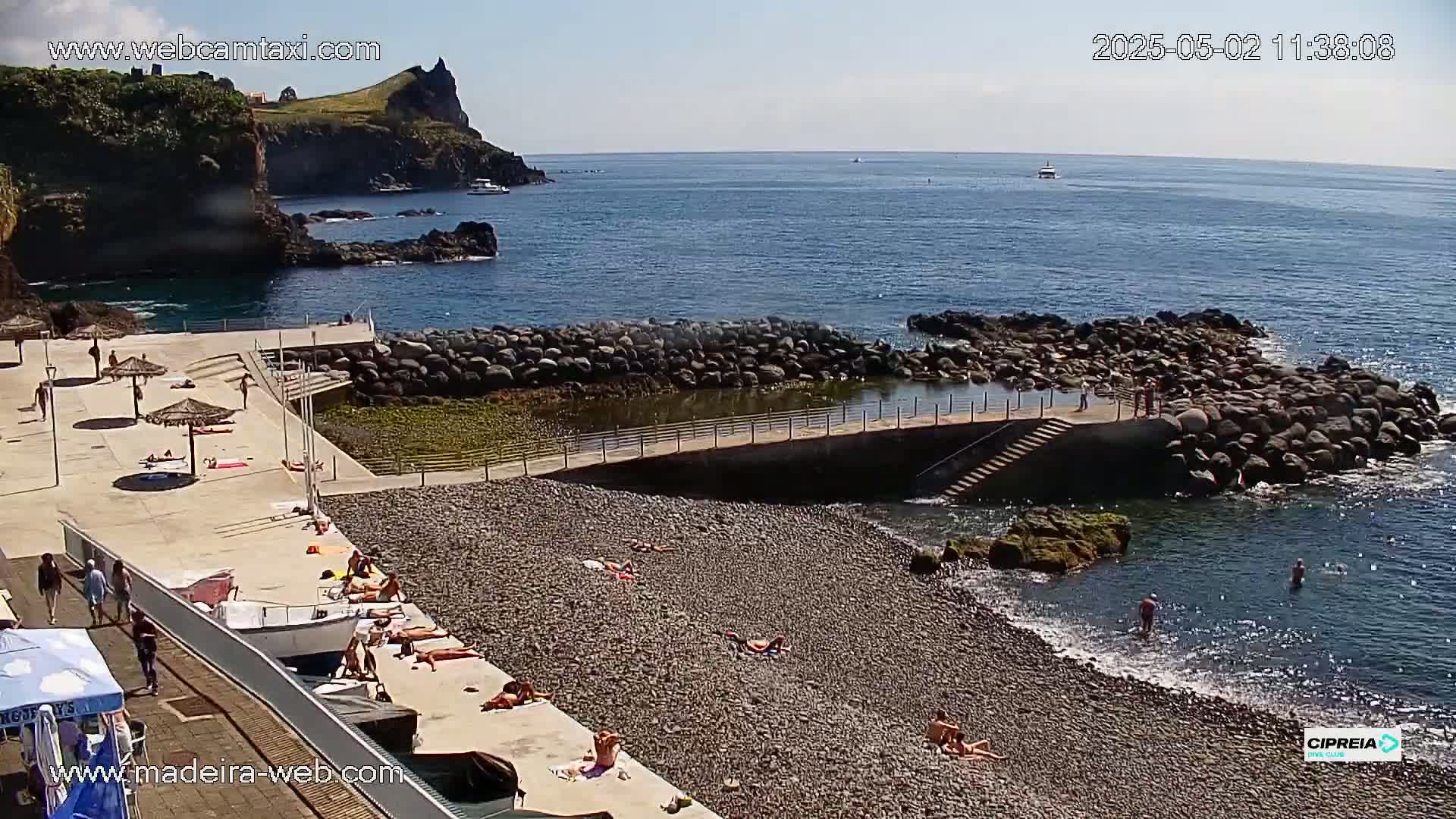 A pebble beach with sunbathers and swimmers, next to a rocky cove and calm ocean under a sunny sky.