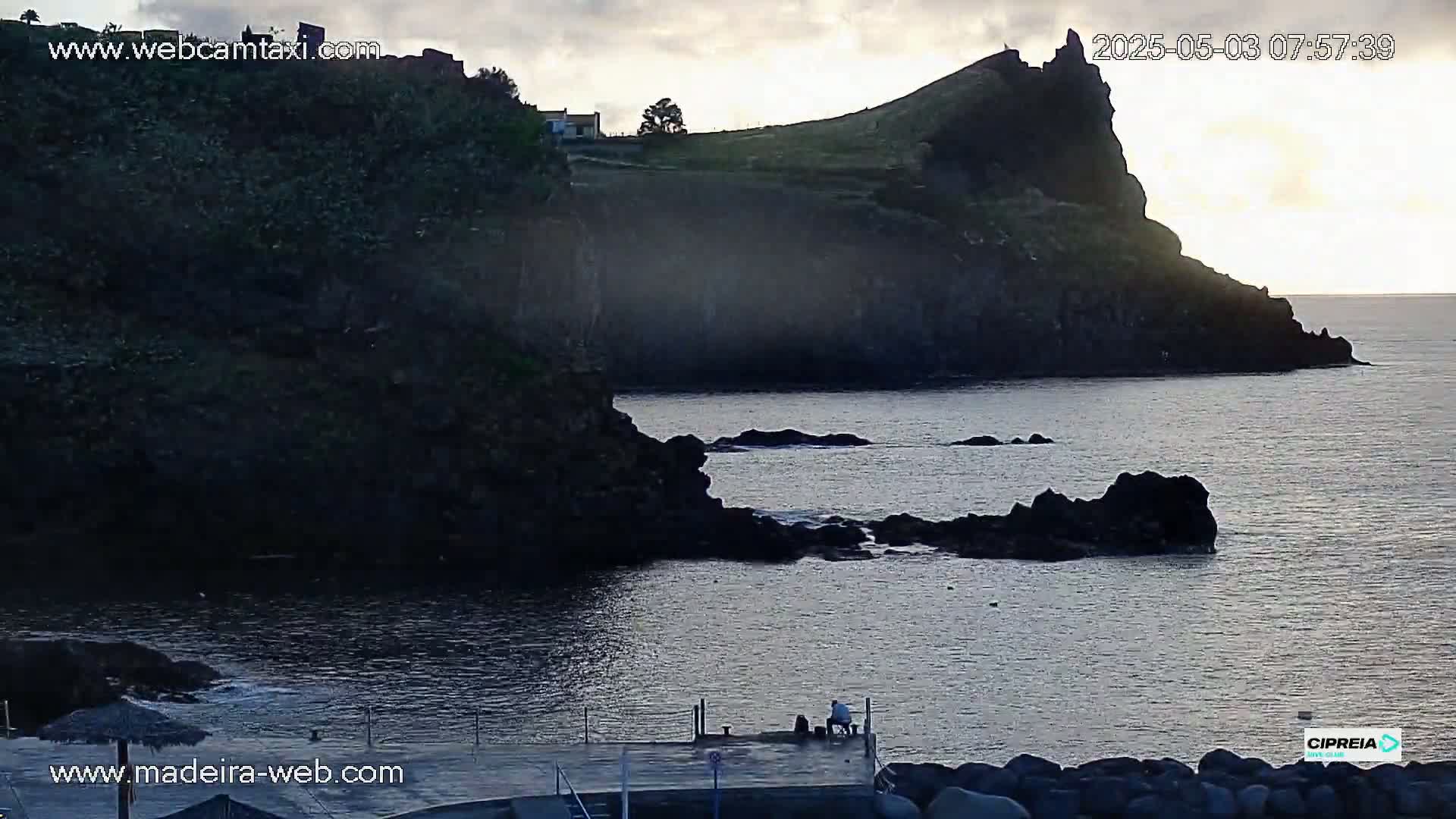 A rocky coastline at sunrise, with dark cliffs and calm water, shows two people sitting on a dock.