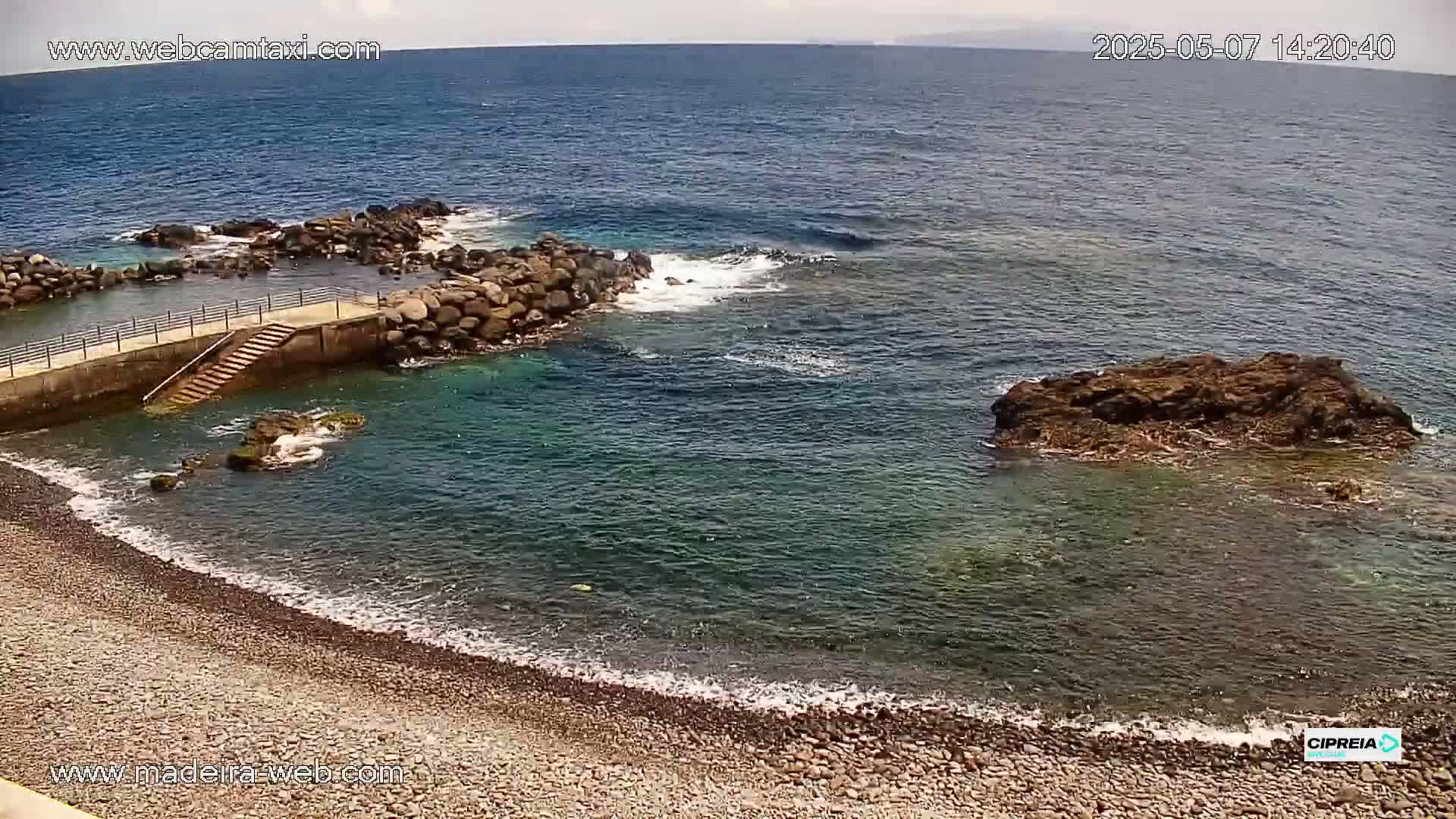 A rocky cove with a small concrete staircase leading to the water, a pebble beach, and calm ocean water under a partly cloudy sky.