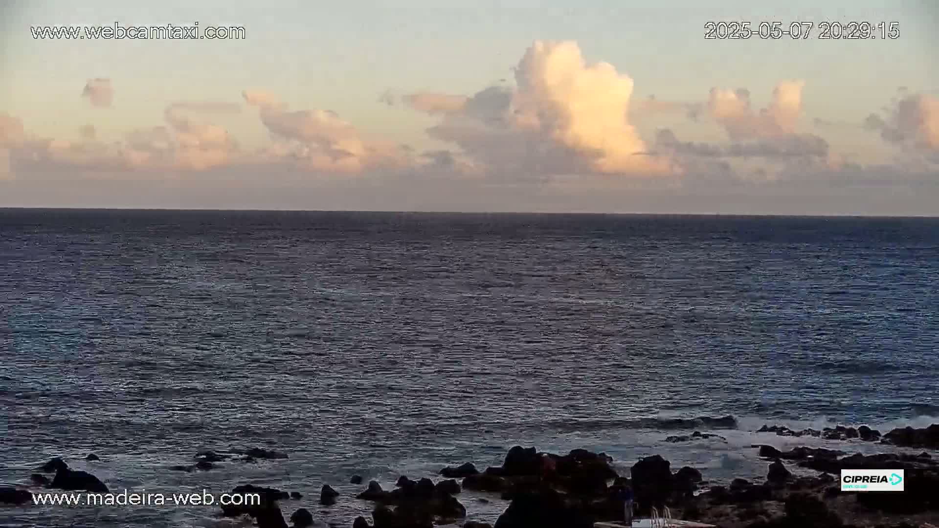 A mostly calm ocean meets a rocky shoreline under a partly cloudy, dusky sky.
