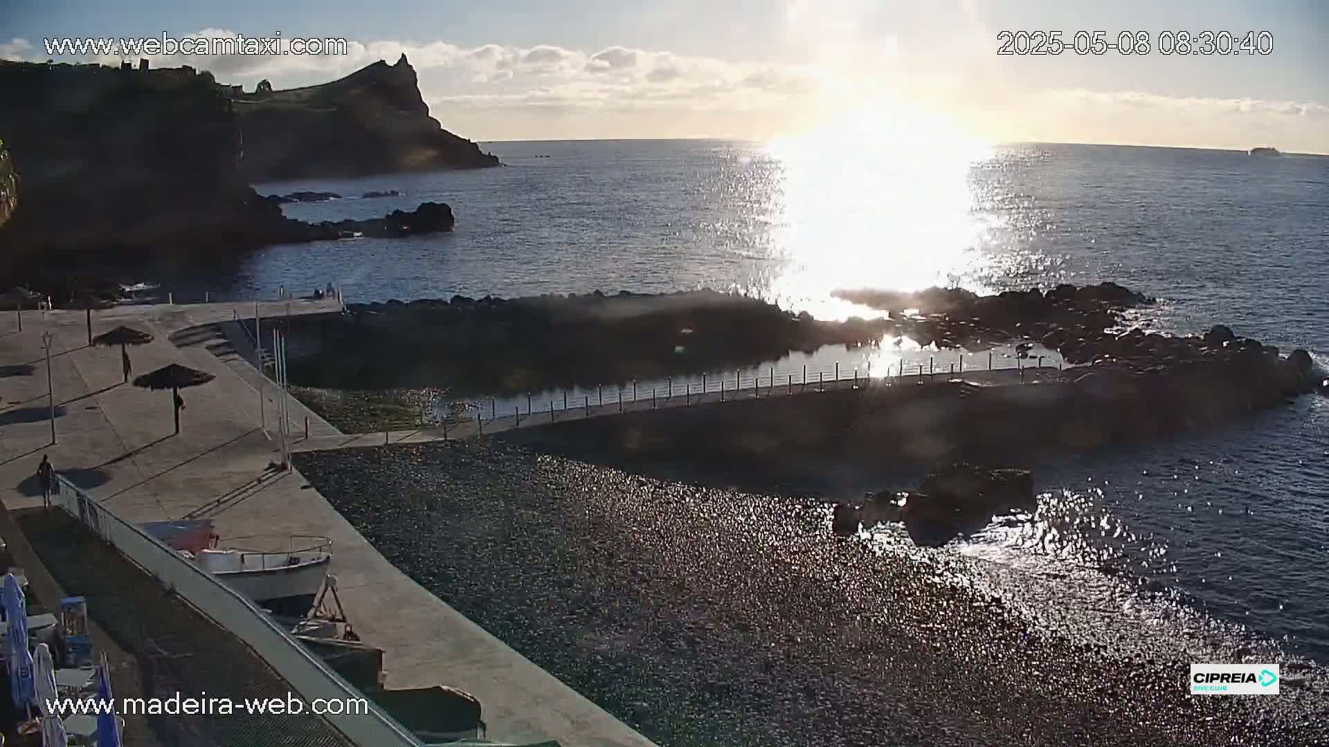 A rocky coastline with a small beach and a walkway leading to a calm pool of water, bathed in the bright sunlight of a clear morning.