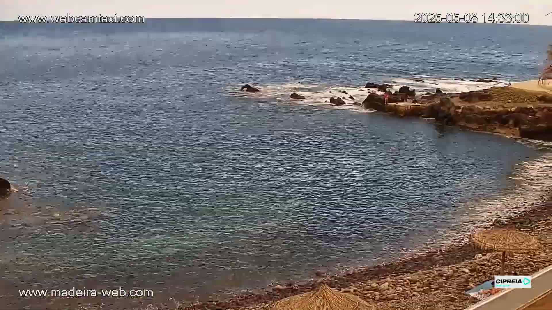 A rocky cove with calm, blue-green water and a few people near the shore on a partly cloudy day.