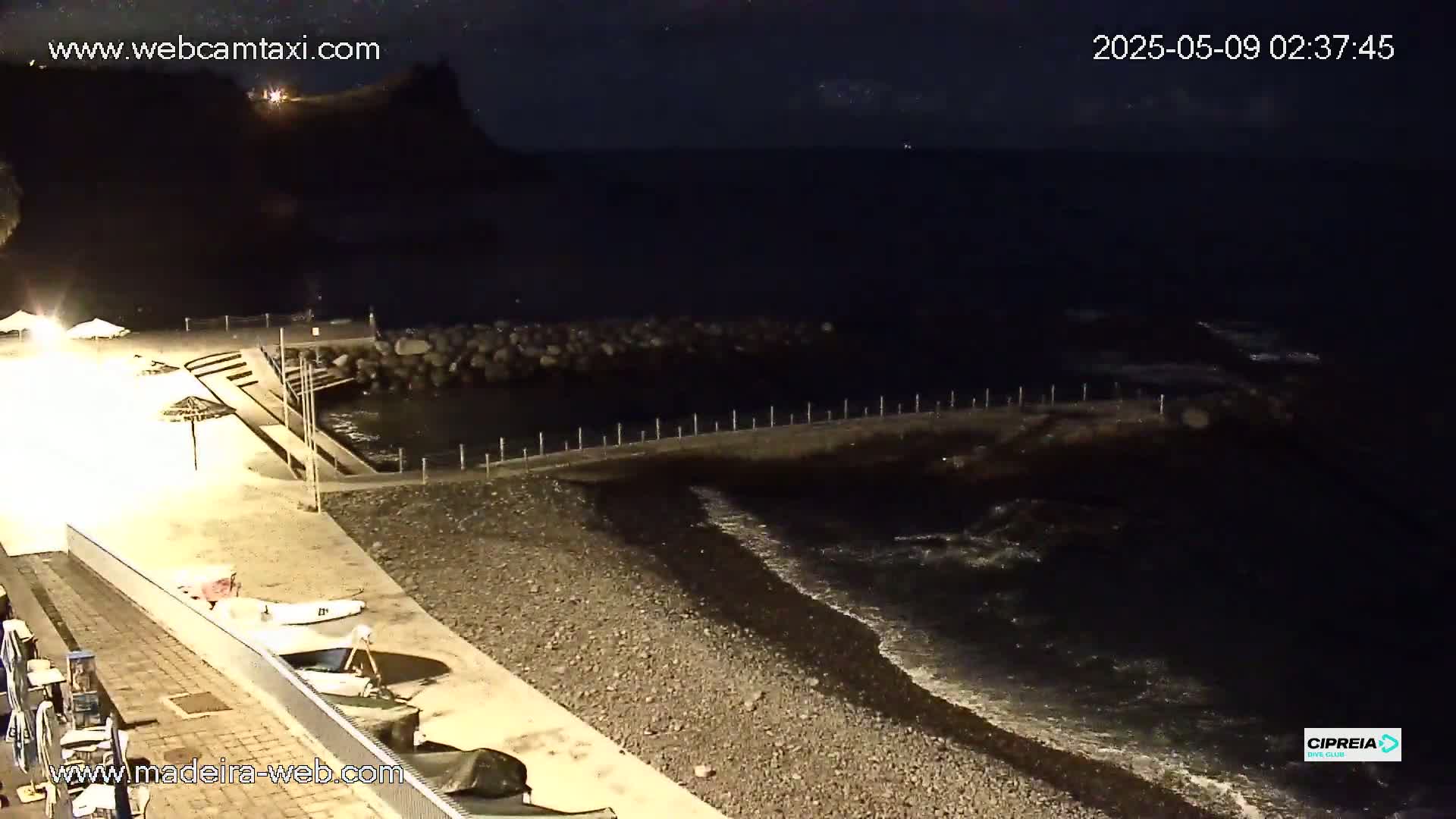A nighttime beach scene shows a dark ocean with gentle waves lapping a pebble shore near a small jetty, with some beach umbrellas and boats visible on the sand.