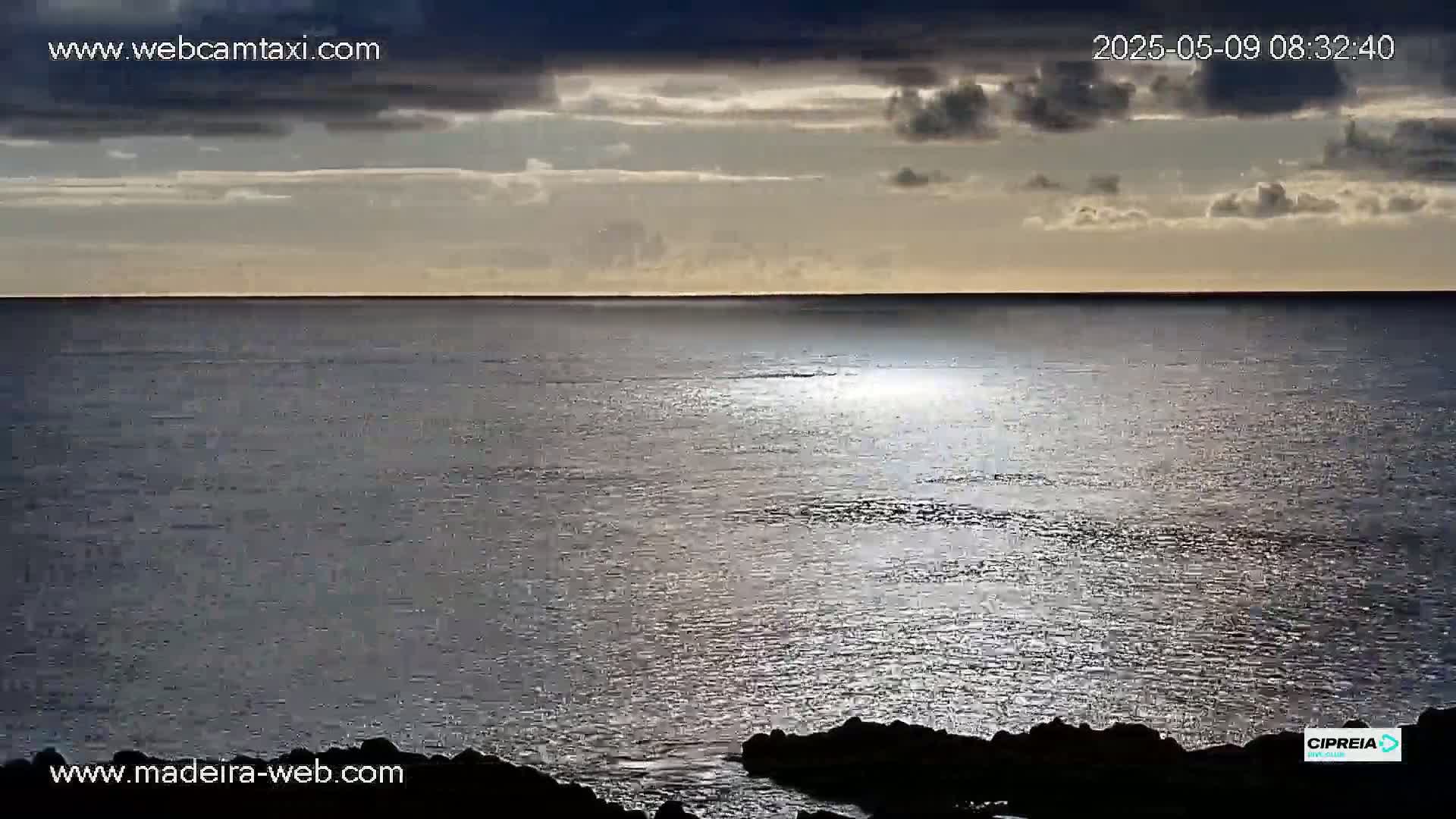 A mostly cloudy sky over a calm, sunlit ocean, with dark rocks visible in the foreground.