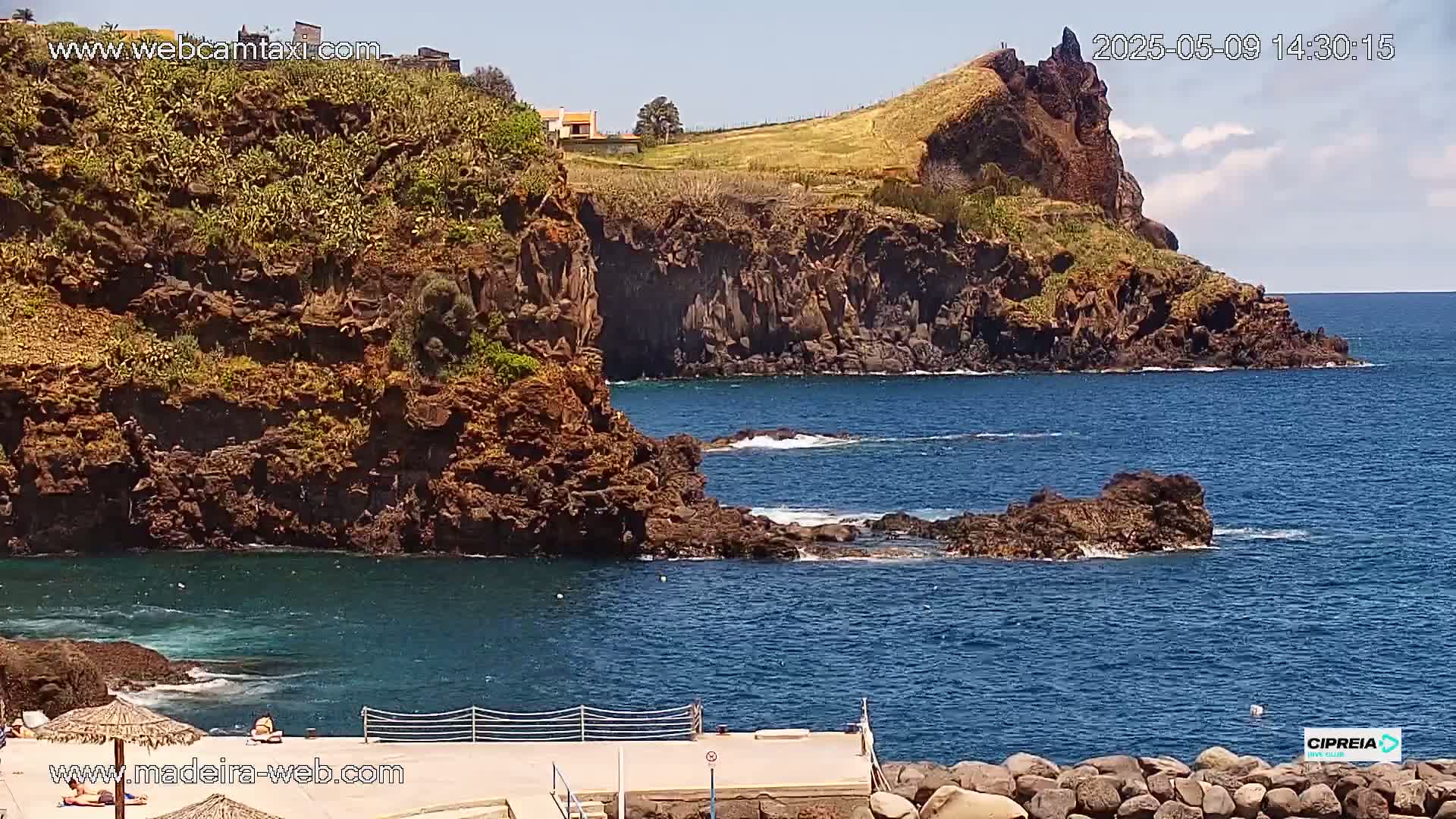 A rocky coastline with dark-colored cliffs and a small, calm cove under a sunny sky.