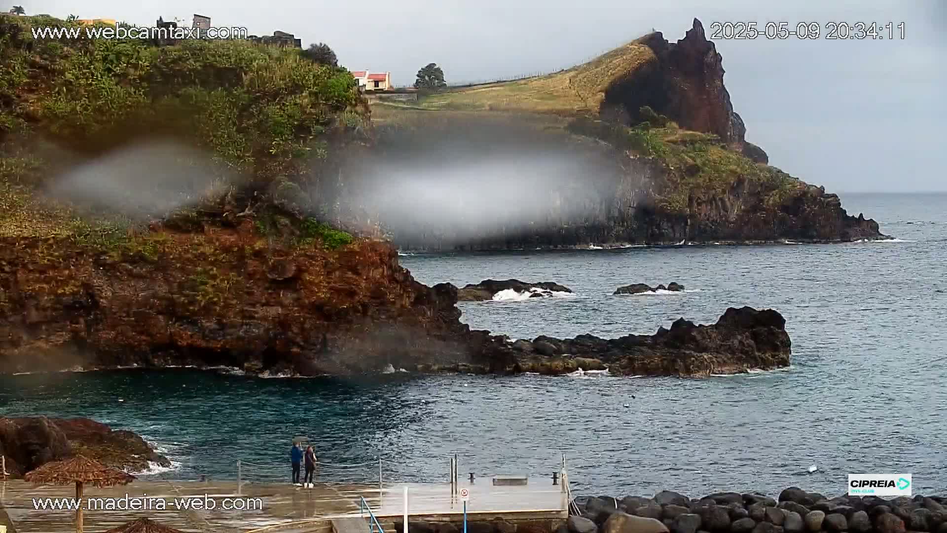 A rainy day reveals a rocky coastline with dark, volcanic-looking cliffs and a small, calm cove where two people stand on a pier.