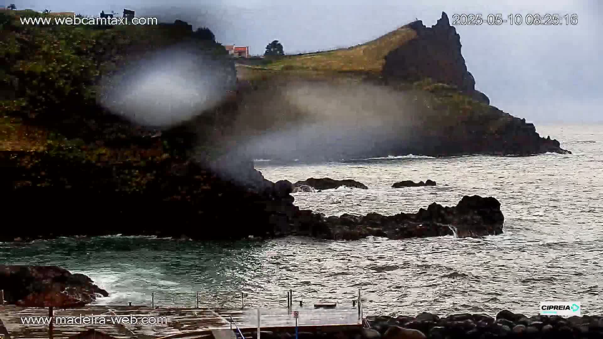 A misty spray of water rises from dark, rocky cliffs into an overcast sky above a churning ocean.