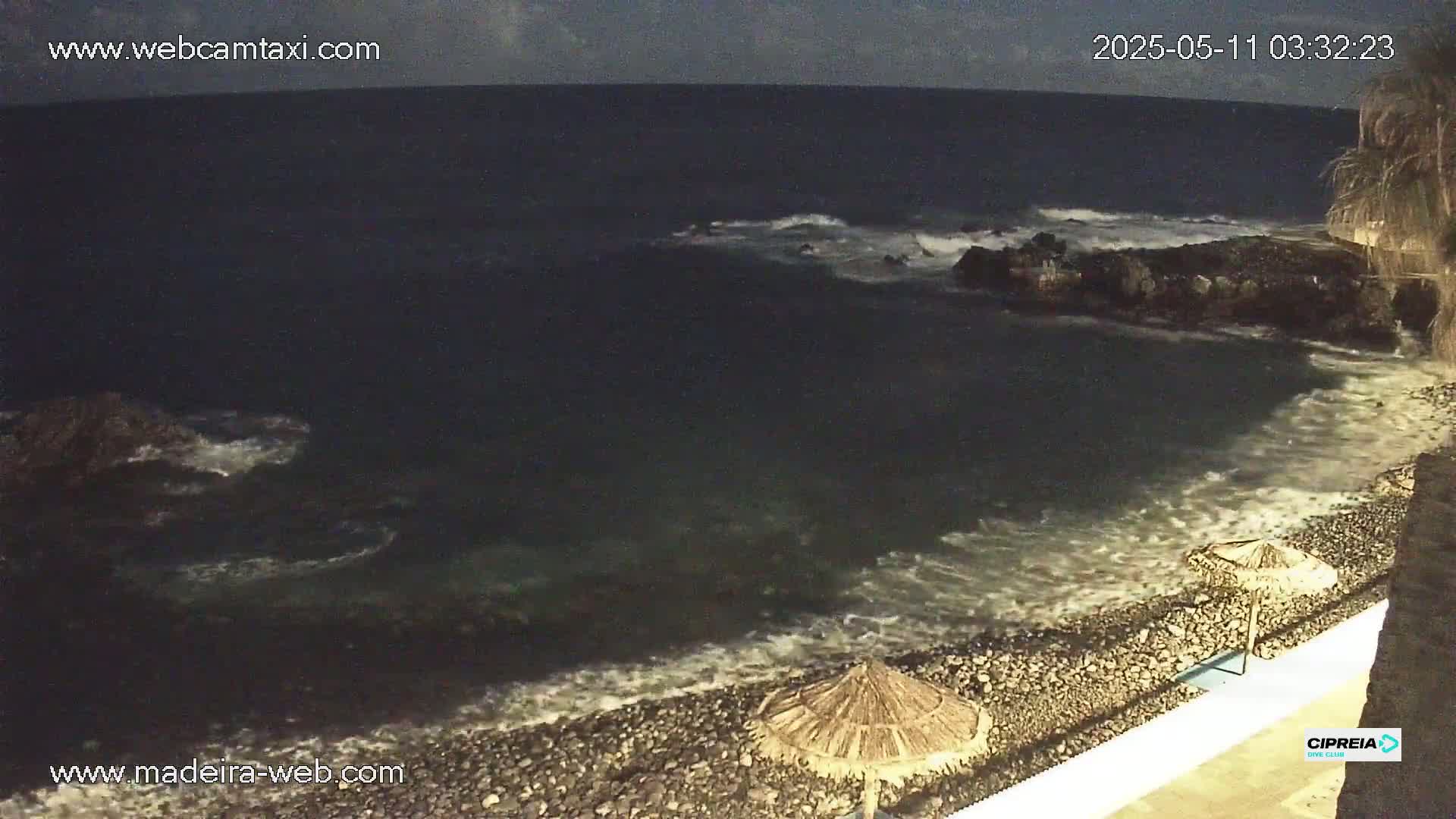 A dark, rocky beach at night is lapped by small waves under a cloudy sky.