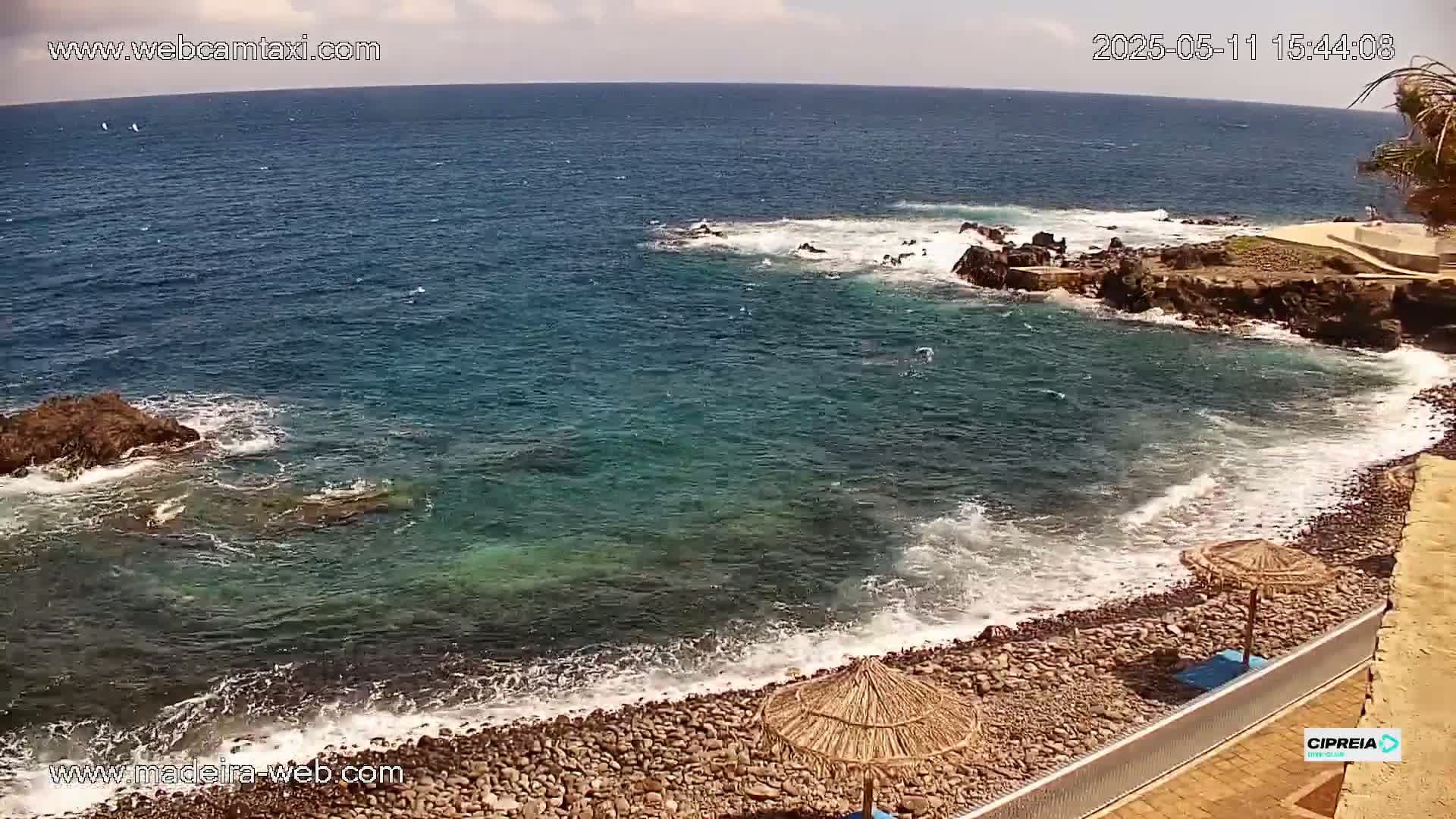 A rocky beach with a few straw umbrellas is lapped by light blue-green ocean waves under a partly cloudy sky.